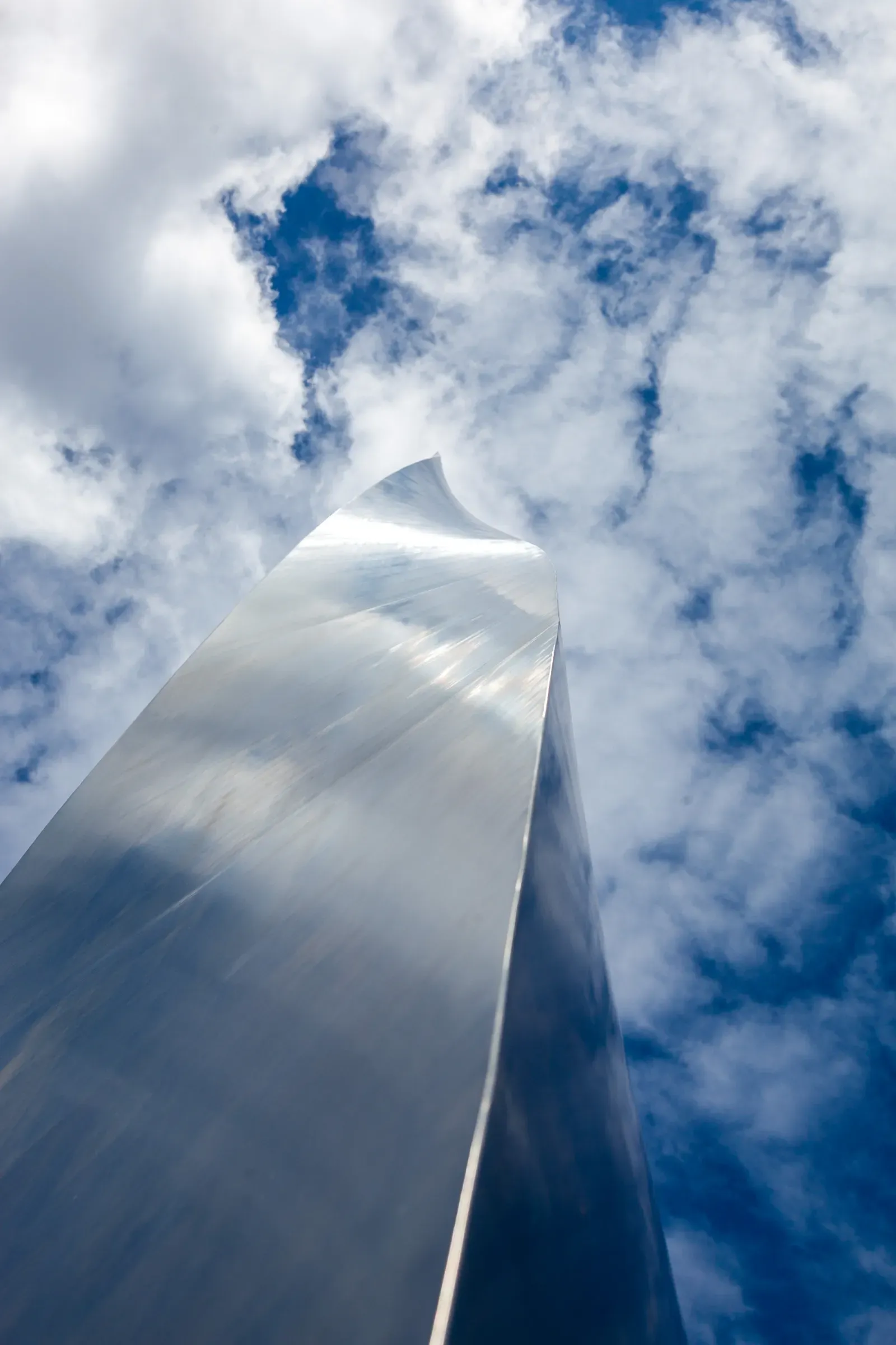 Looking straight up along the folded steel edge of a tall abstract sculpture against a blue sky with clouds