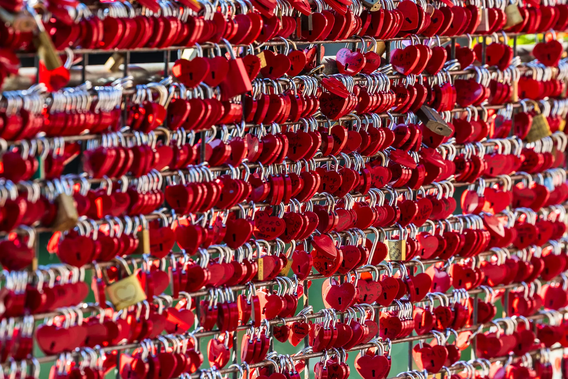 Rows of red heart-shaped padlocks covering a bridge railing, backlit by the sun.