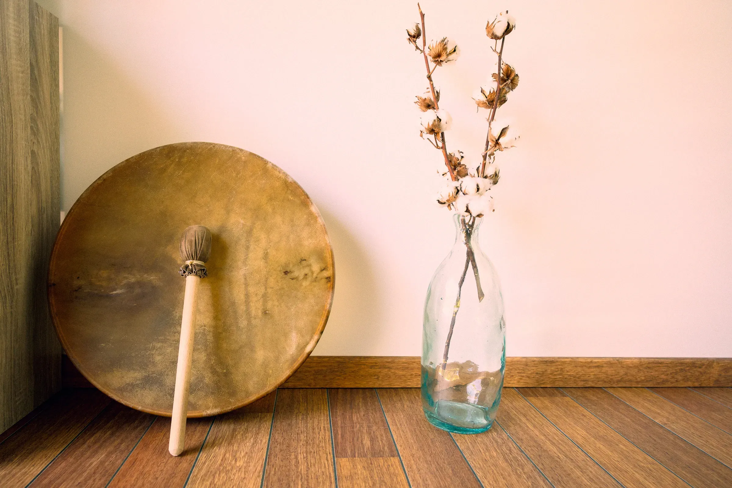 Frame drum with a round mallet resting across it, leaning against a cream wall beside a clear glass bottle holding dried cotton branches, warm light on a wooden floor