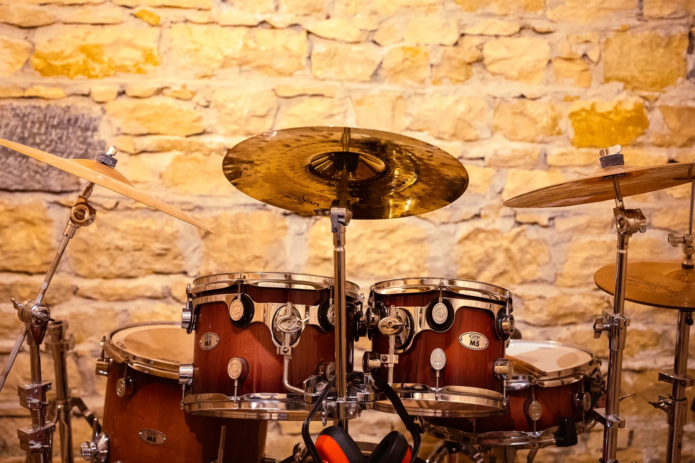 Full PDP M5 drum kit with a gold Alchemy cymbal overhead, orange headphones resting on the bass drum, warm stone wall behind, Lyon