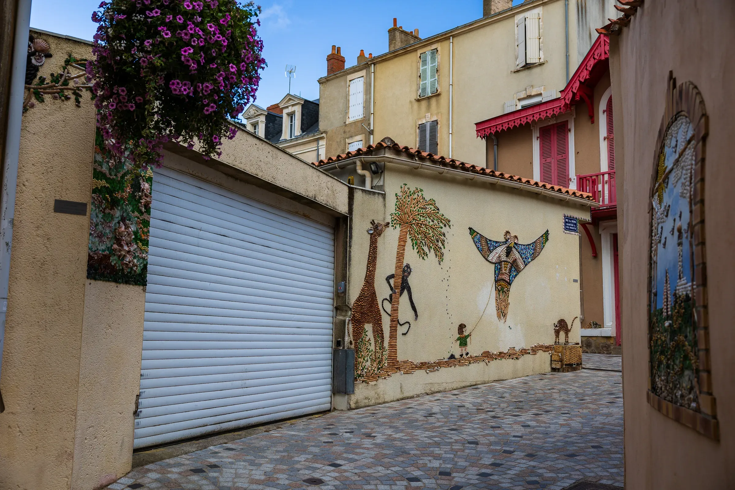 Wide cobblestone alley with a shell mosaic mural of giraffes, a climbing figure, and birds on a garage wall