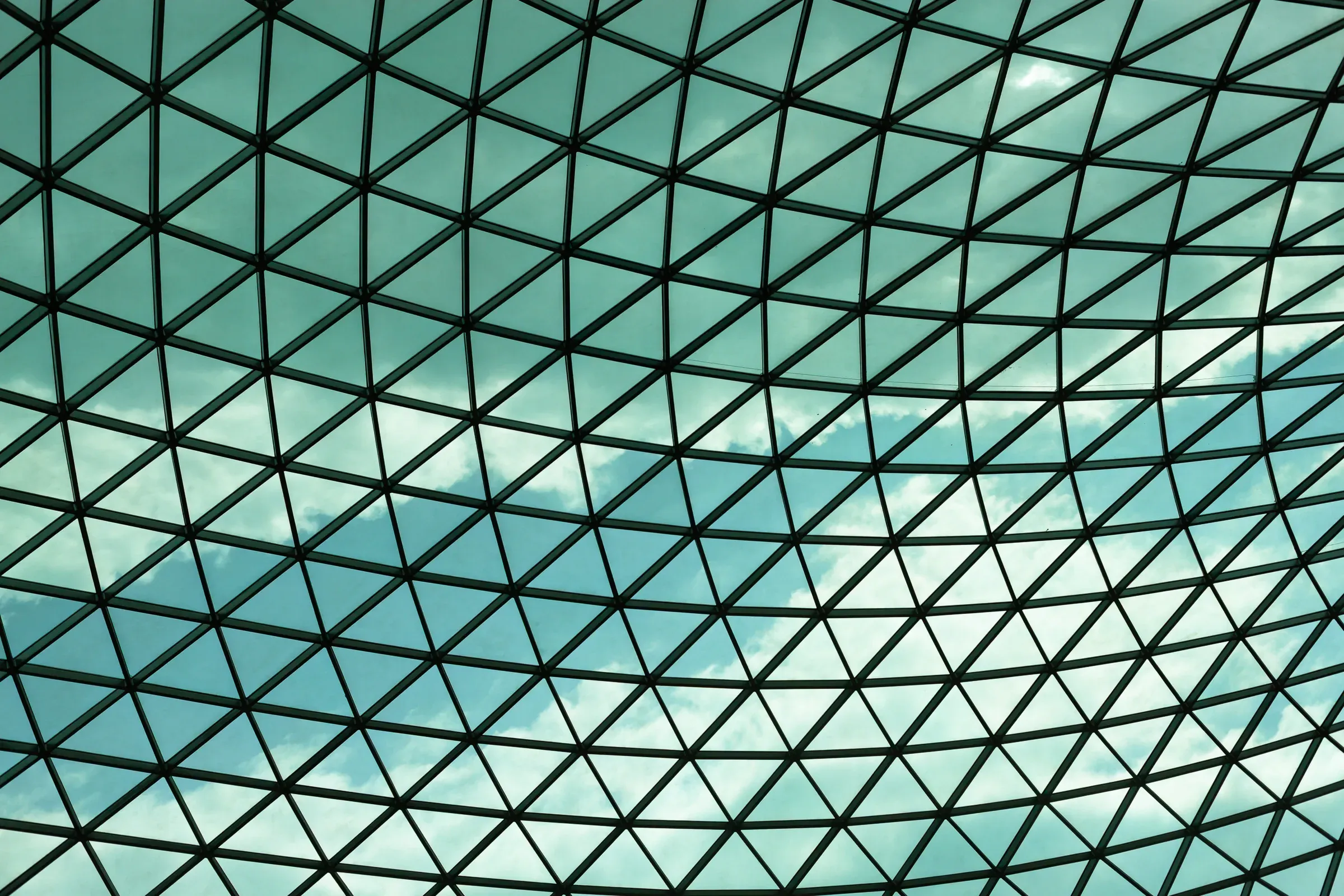 Triangulated glass and steel roof of a museum courtyard, clouds visible through the panels
