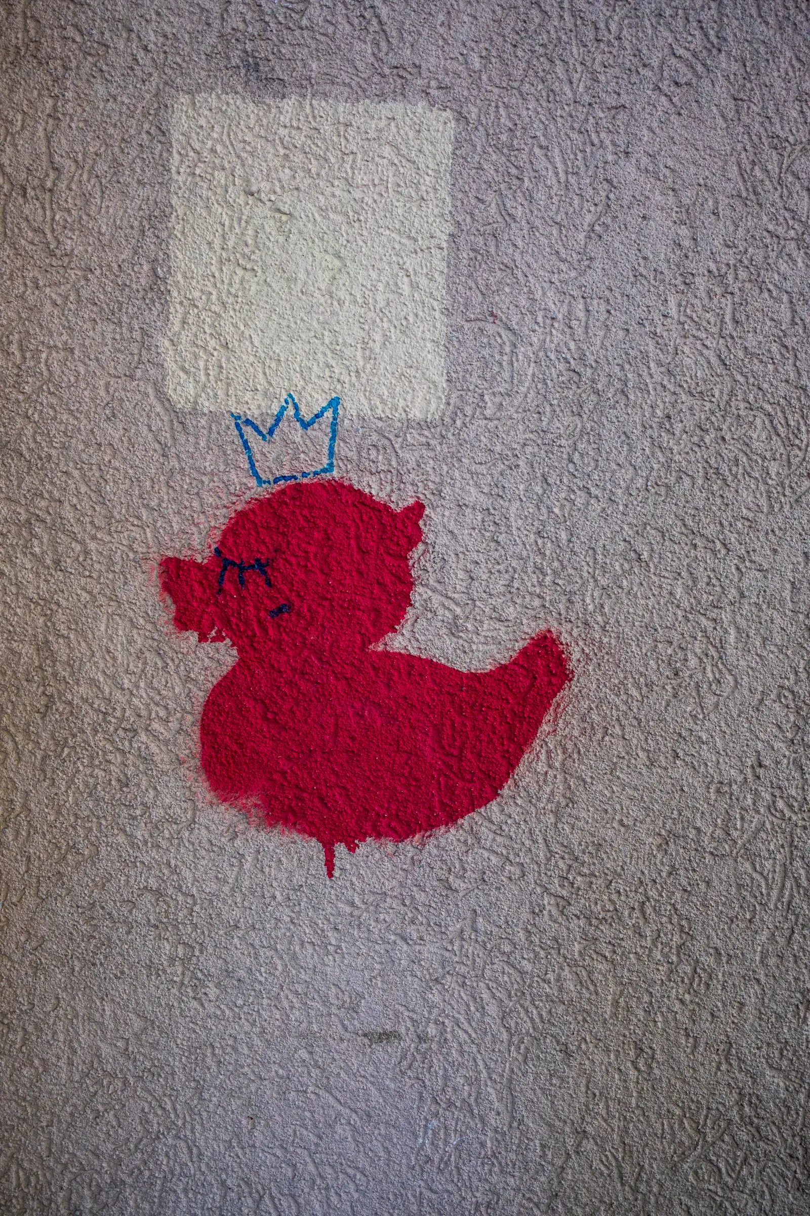 A red rubber-duck stencil wearing a blue crown, painted on a stucco wall.
