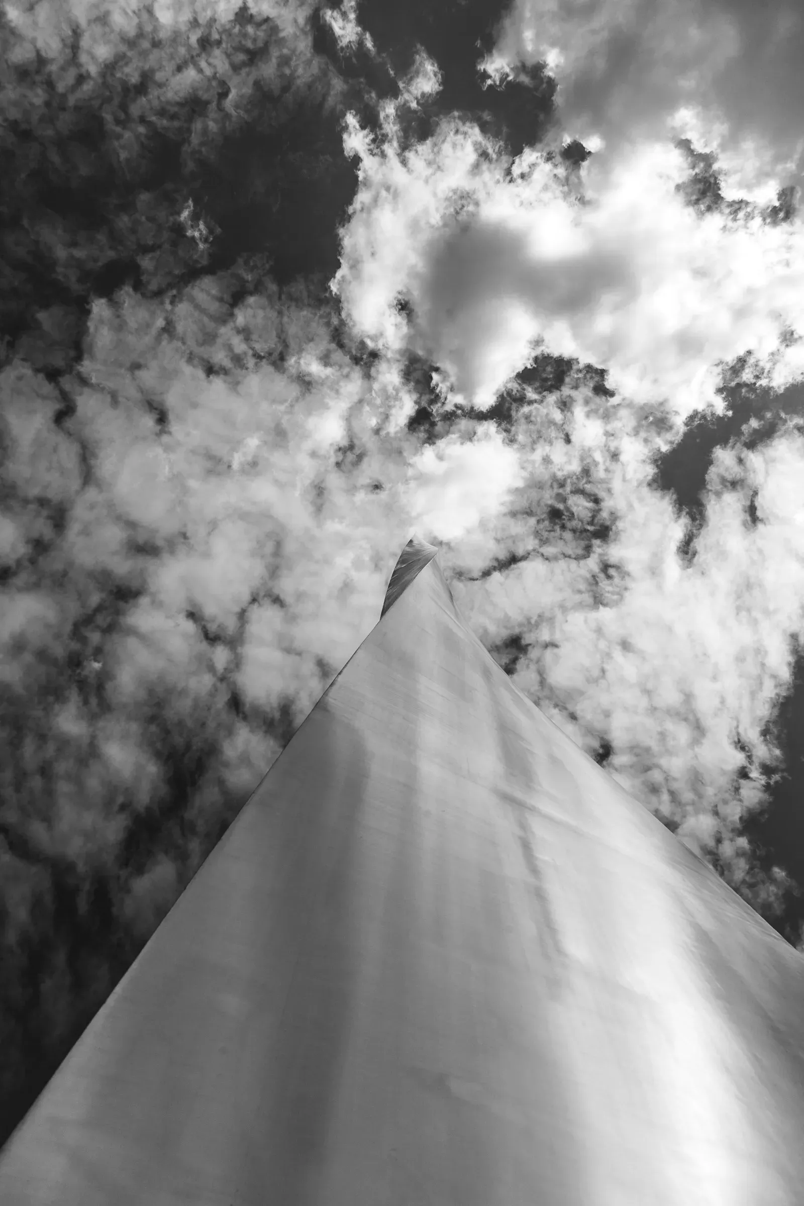 Looking straight up at a building's sharp glass corner against a cloud-filled sky, black and white