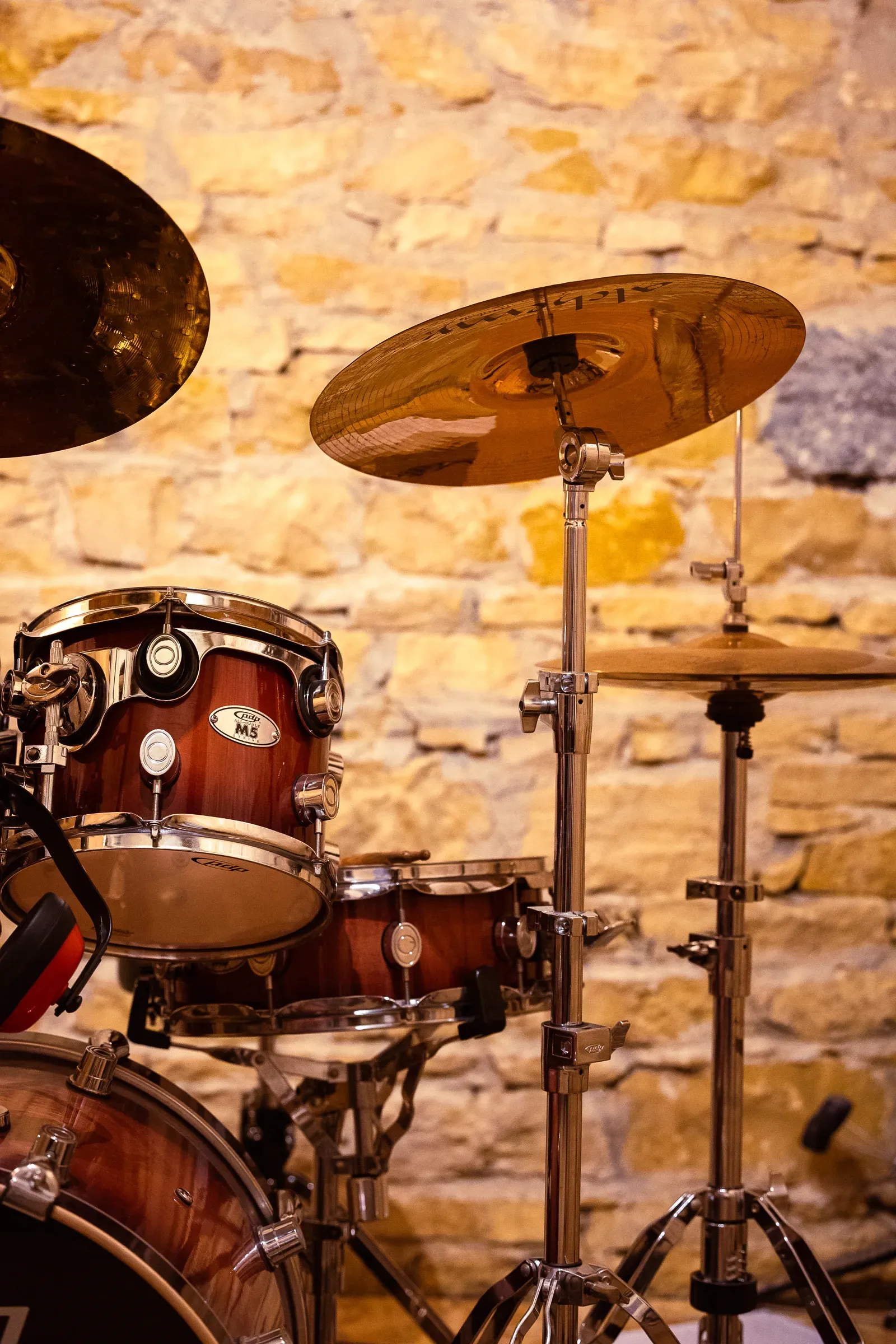 Side view of the drum kit — snare, toms, and two cymbals against a warm stone wall, Lyon