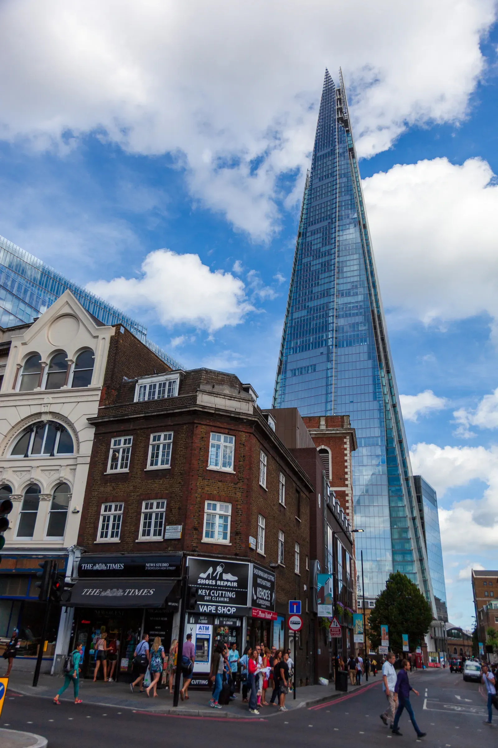 The Shard rising behind a red-brick corner building with The Times news studio and a shoe repair shop, pedestrians crossing below
