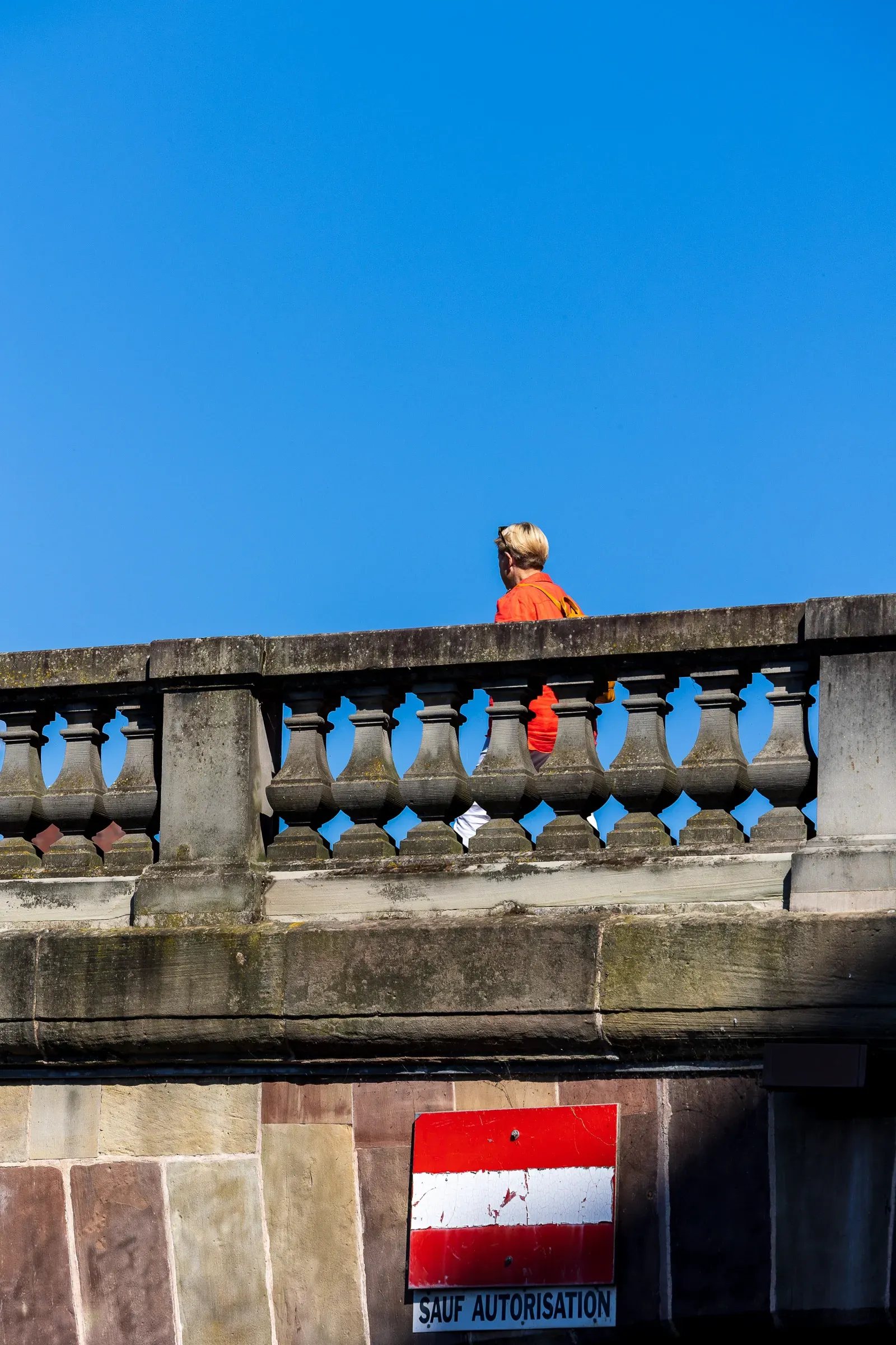 A woman in a red jacket looking out from a stone bridge parapet against a clear sky.