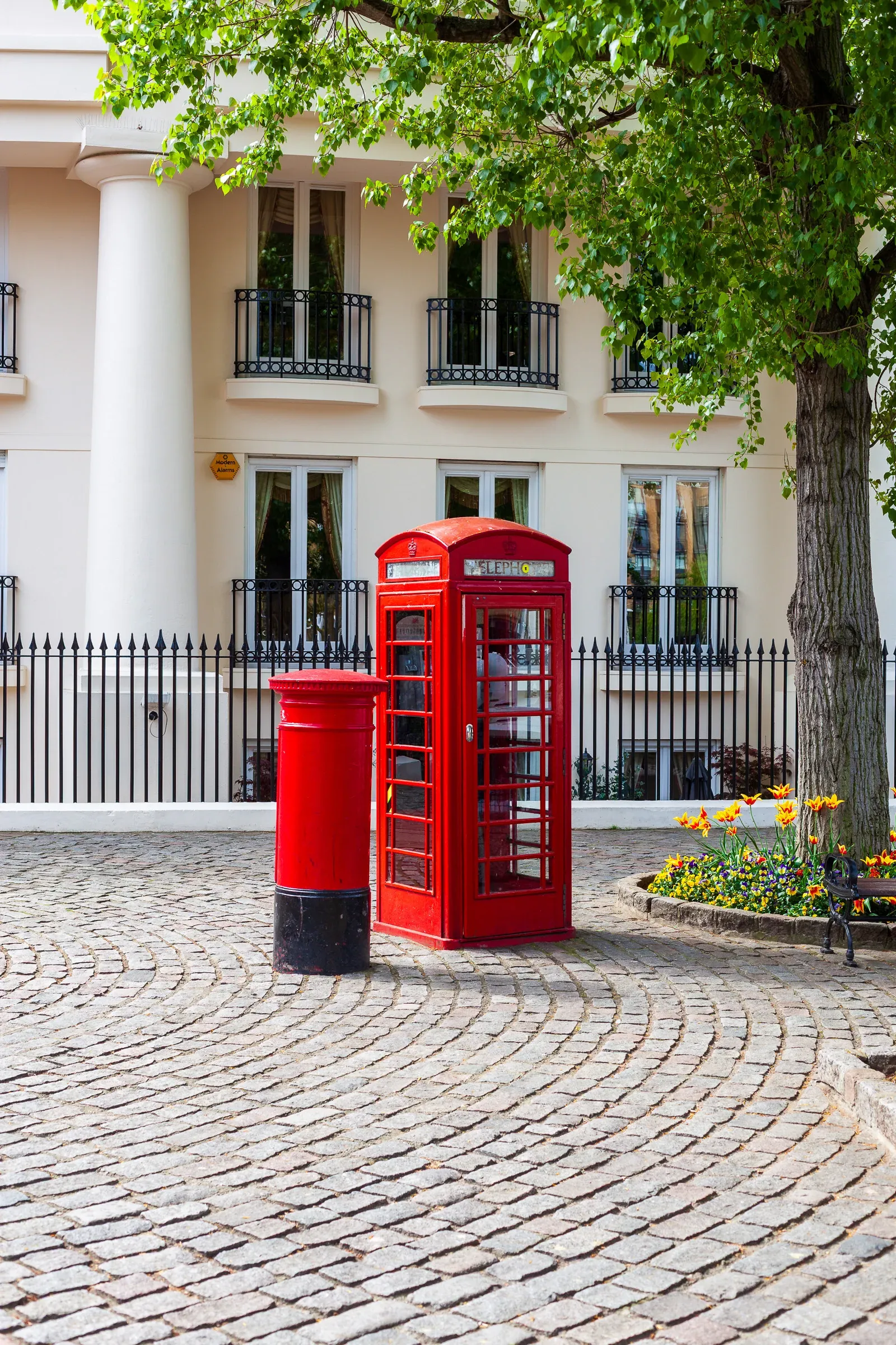 A red pillar box and red telephone box side by side on a cobblestone courtyard, white Georgian building with iron railings behind, a leafy tree to the right