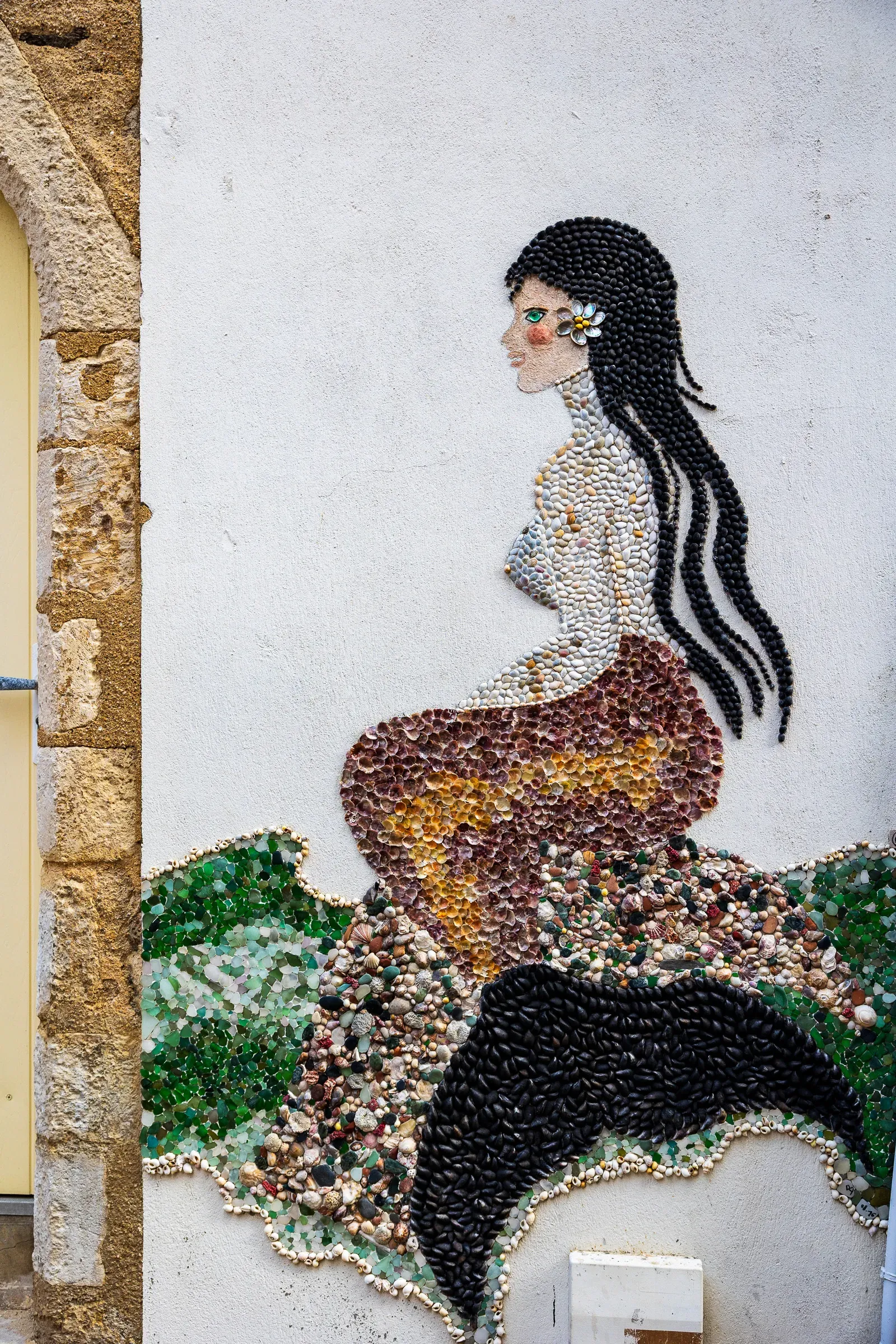 Mermaid figure made of shells in profile on a white wall beside a stone doorframe