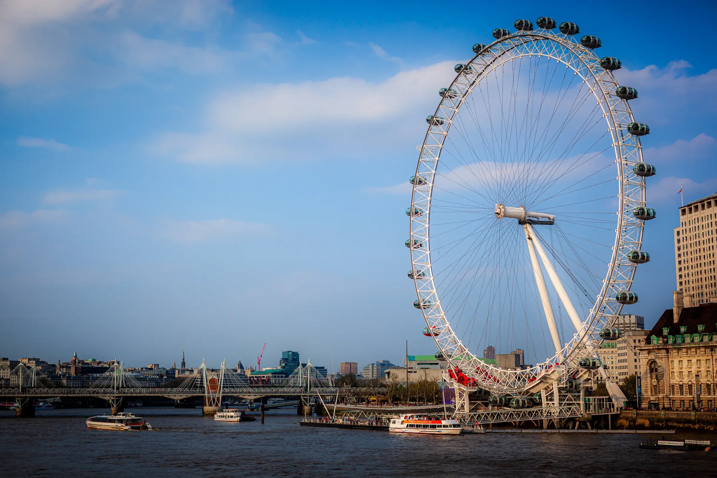The London Eye observation wheel against blue sky, the Thames below with tour boats and Hungerford Bridge stretching into the distance