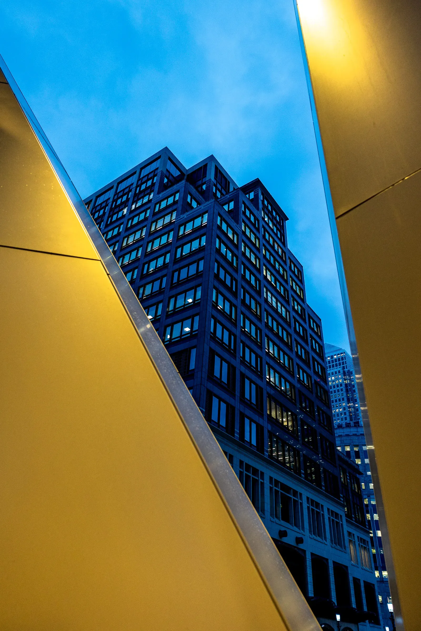 An office tower at dusk framed between two mustard-yellow angled walls, blue sky above