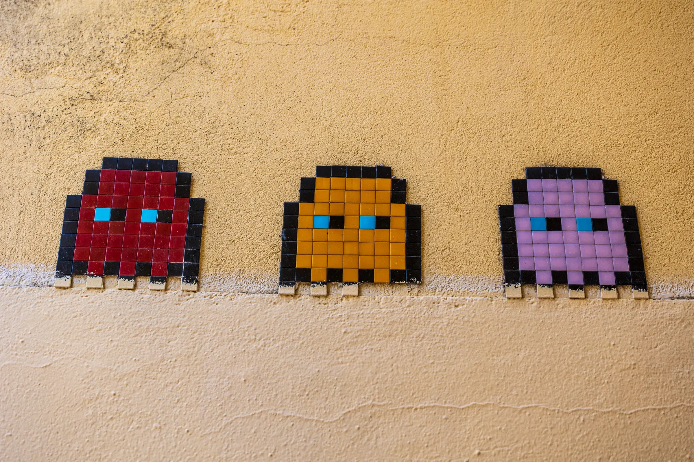 Three pixel-art mosaic ghosts, red, orange, and purple, set into a mustard-yellow wall.