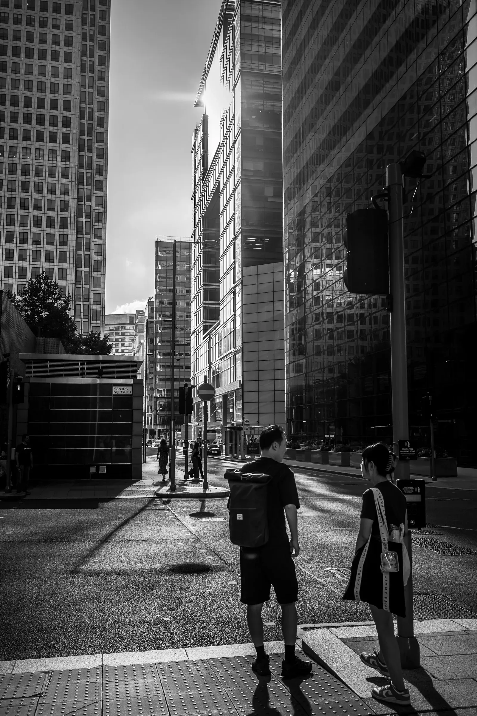 Two pedestrians waiting to cross a street between glass office towers near Canada Square, sun flare above, black and white