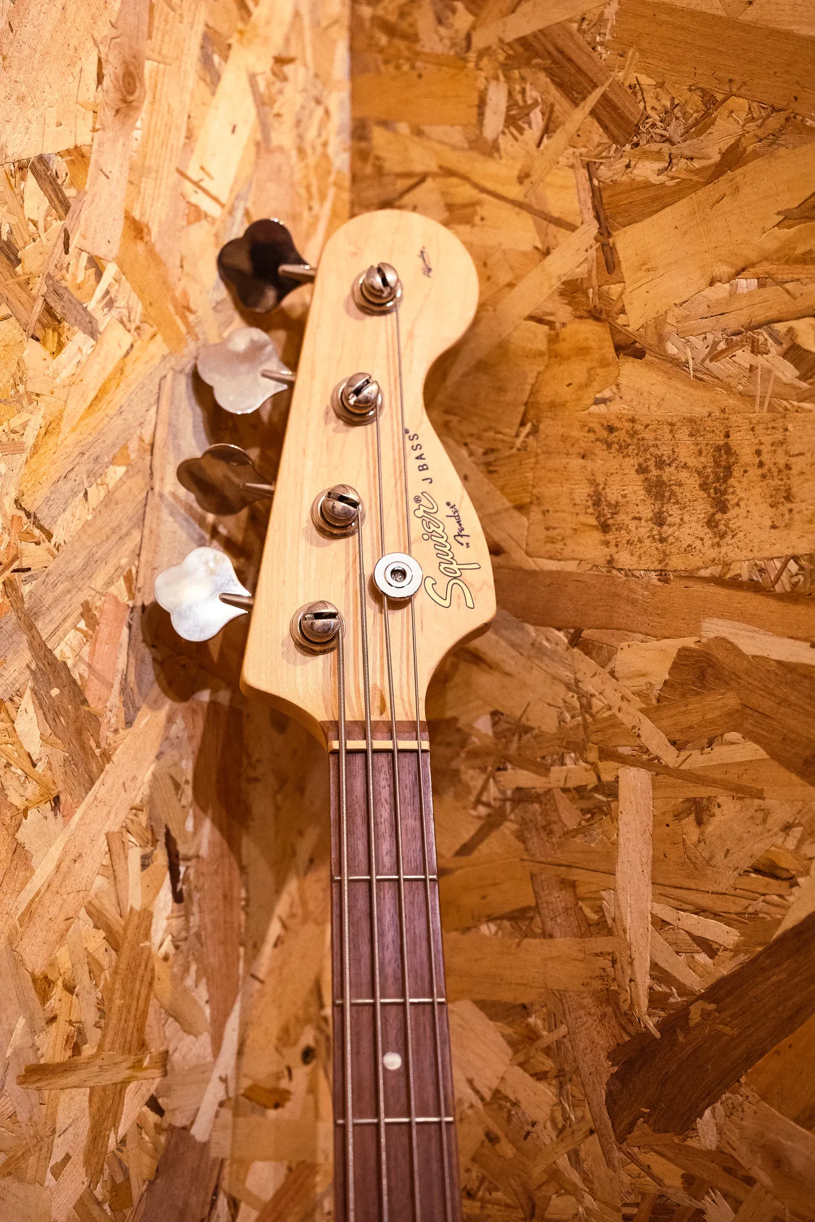 Squier Jazz Bass headstock leaning against an OSB wall, tuning pegs and strings visible, Lyon