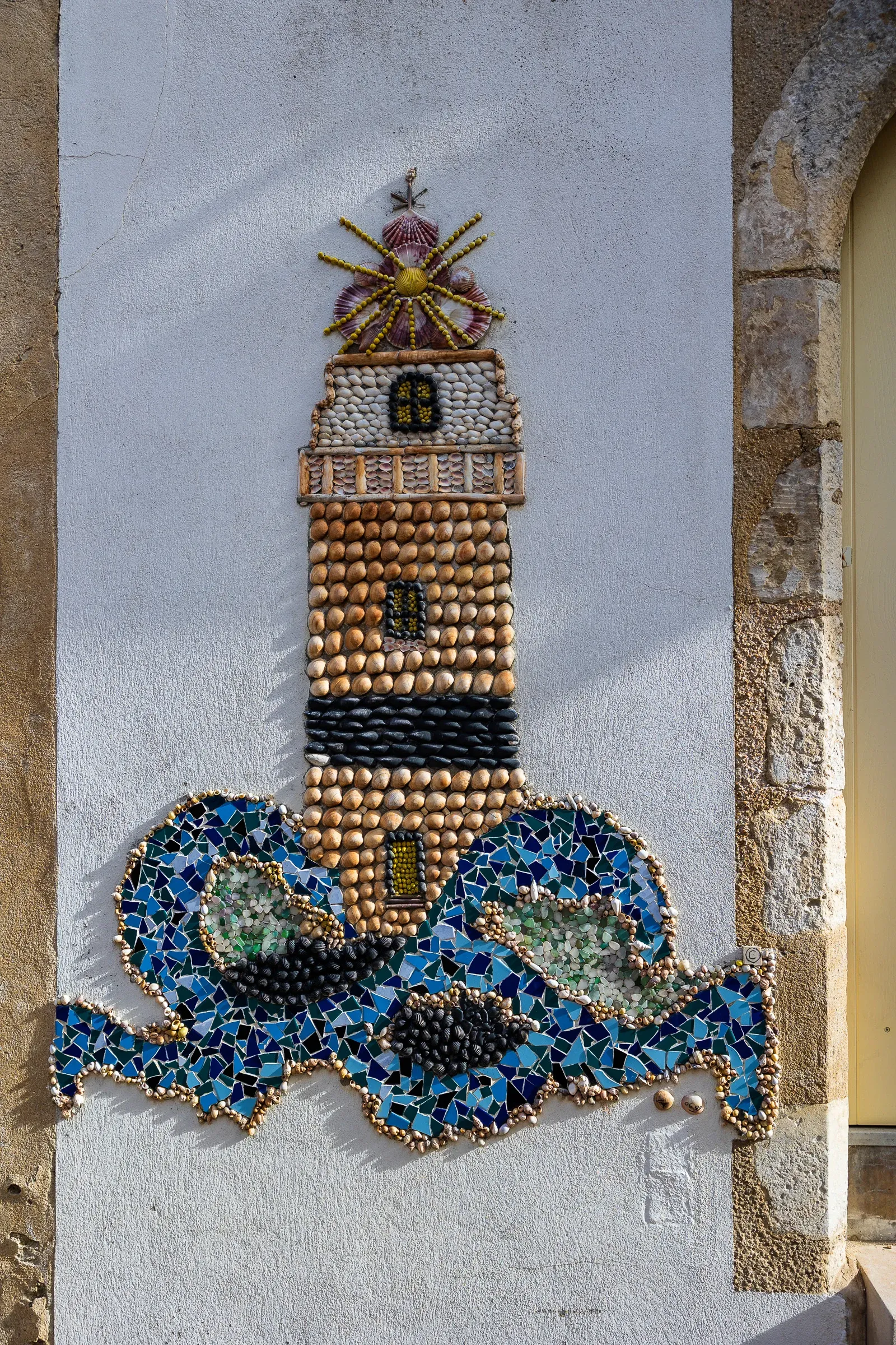 Lighthouse made of shells and mosaic tiles on a white wall framed by dressed stone blocks