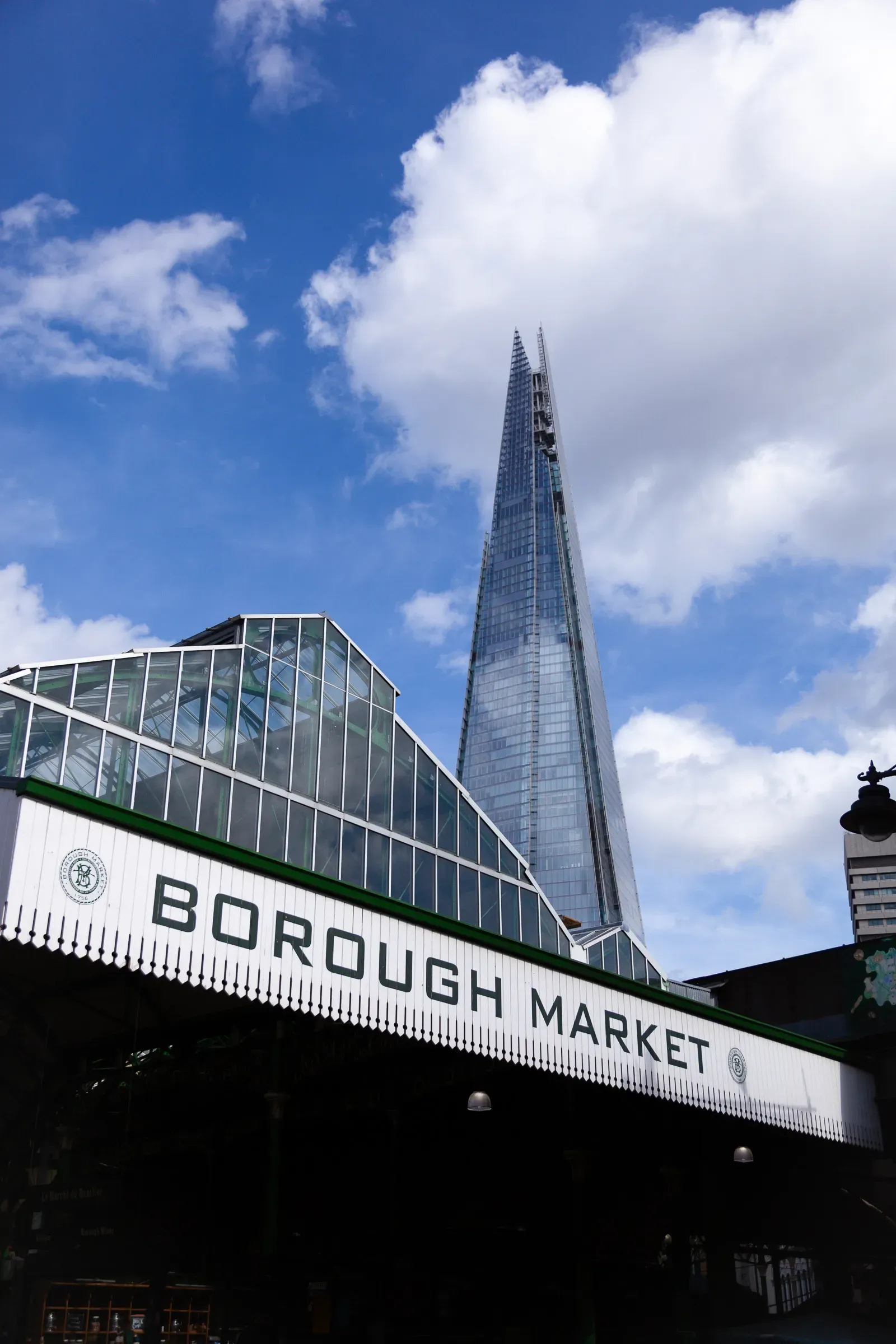 Borough Market entrance sign with The Shard rising sharply behind it, blue sky with scattered white clouds