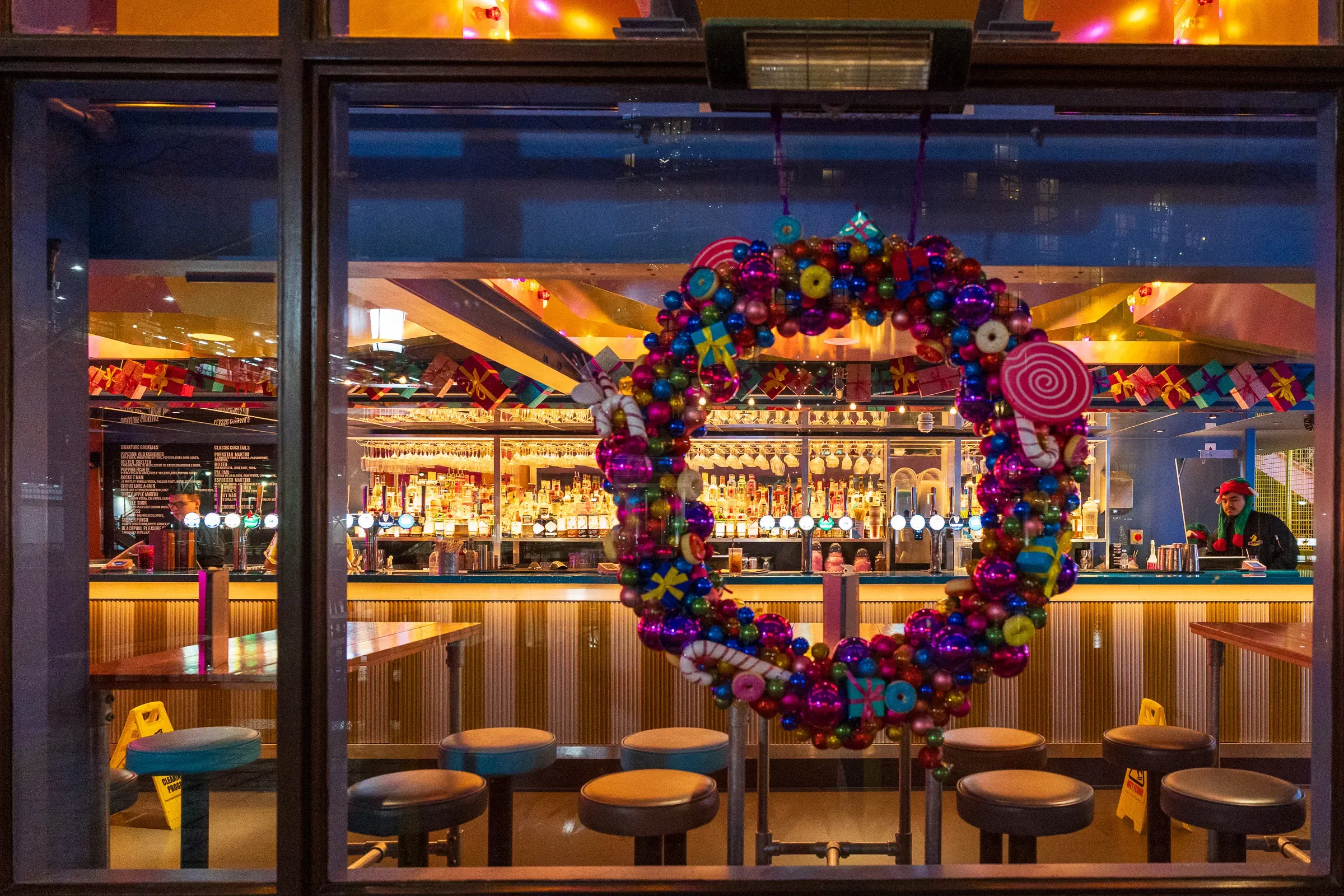A Christmas wreath of baubles and candy canes hanging in a bar window, the bar and its bottles lit up behind