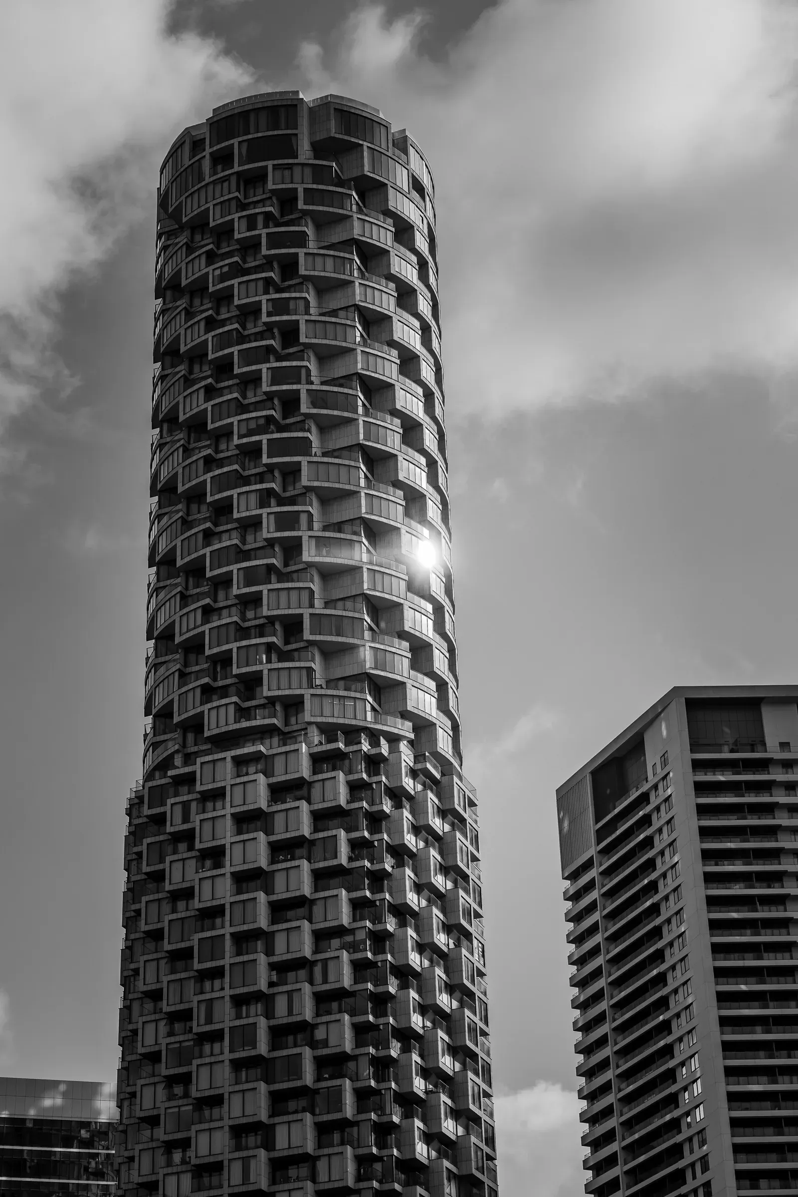 Low angle view of a tall curved residential tower against a cloudy sky, a second high-rise beside it, black and white