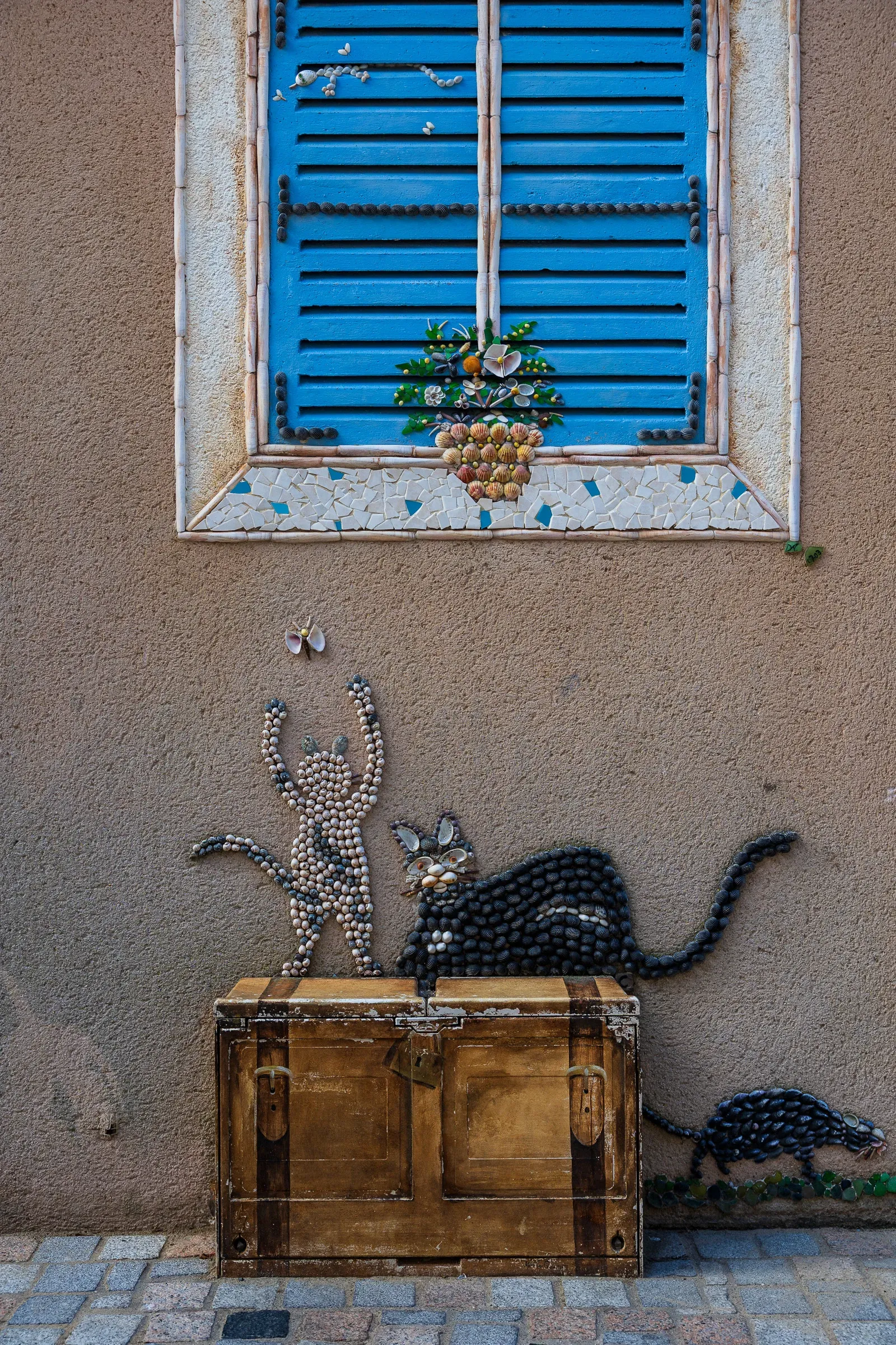 Two shell-art cats playing on a wooden chest below a blue-shuttered window, a third cat on the ground