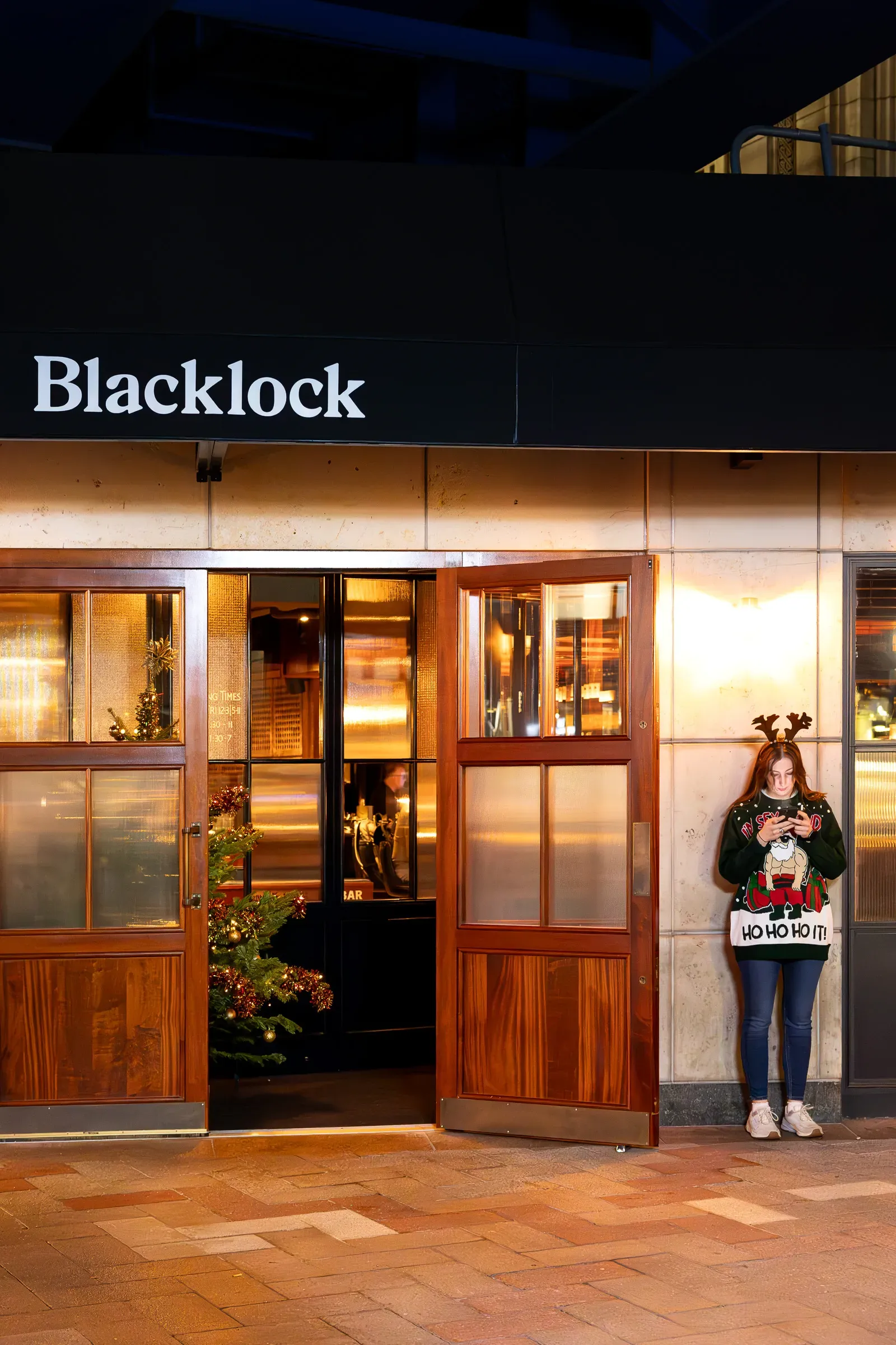 A woman in a Christmas jumper and reindeer antlers checks her phone outside Blacklock restaurant at night, a lit Christmas tree in the doorway