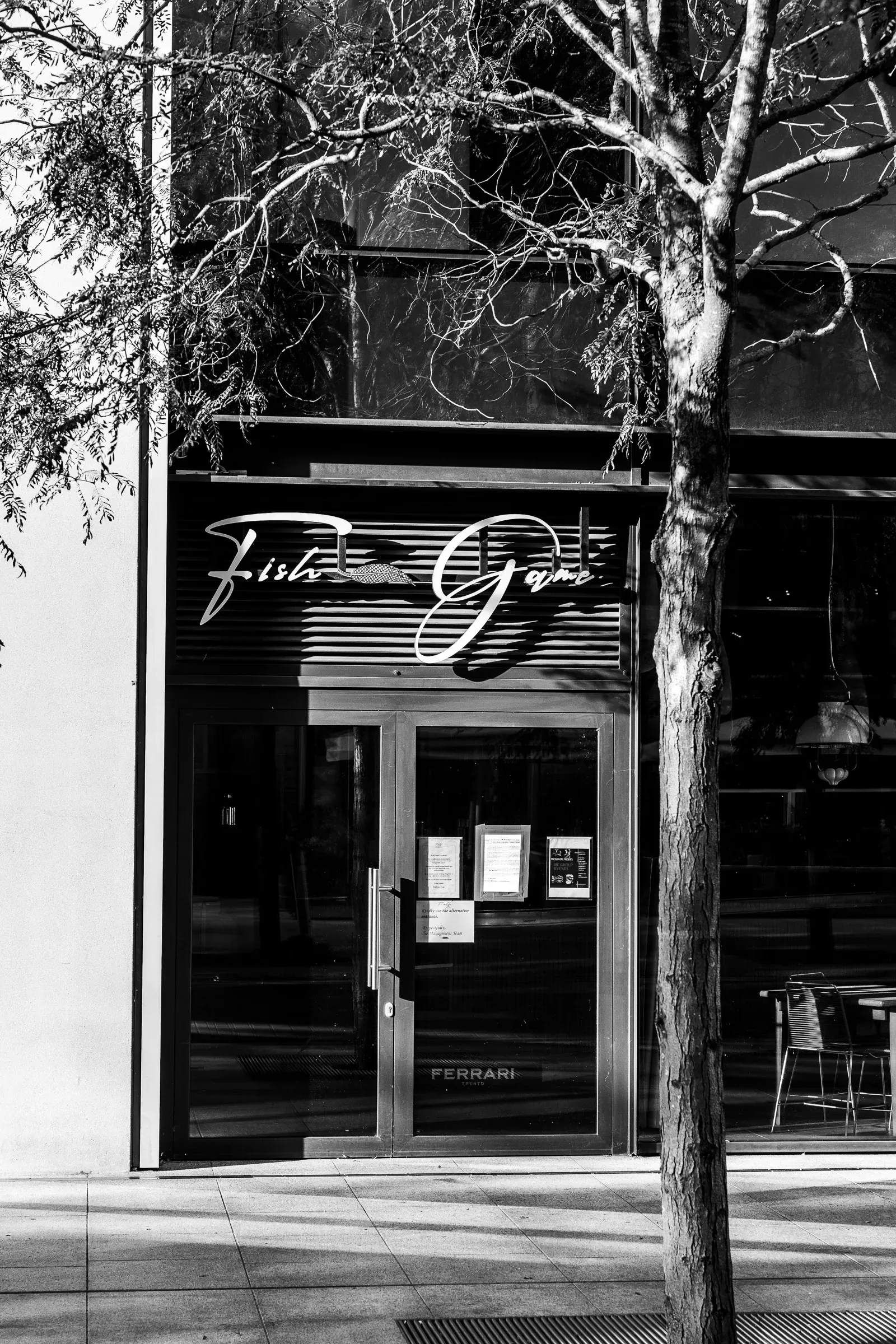 Fish + Game restaurant storefront behind a bare tree, menus posted on the glass door, black and white