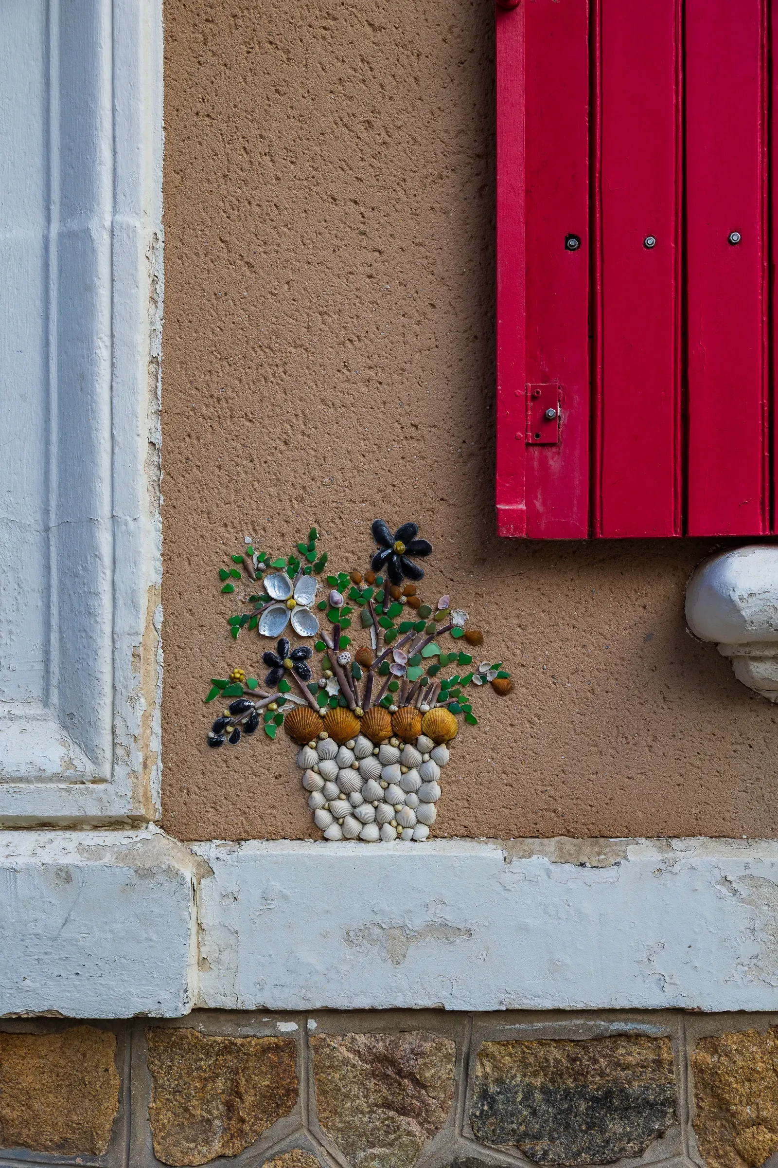 Shell-art flower bouquet in a pot tucked between a white window frame and red shutters