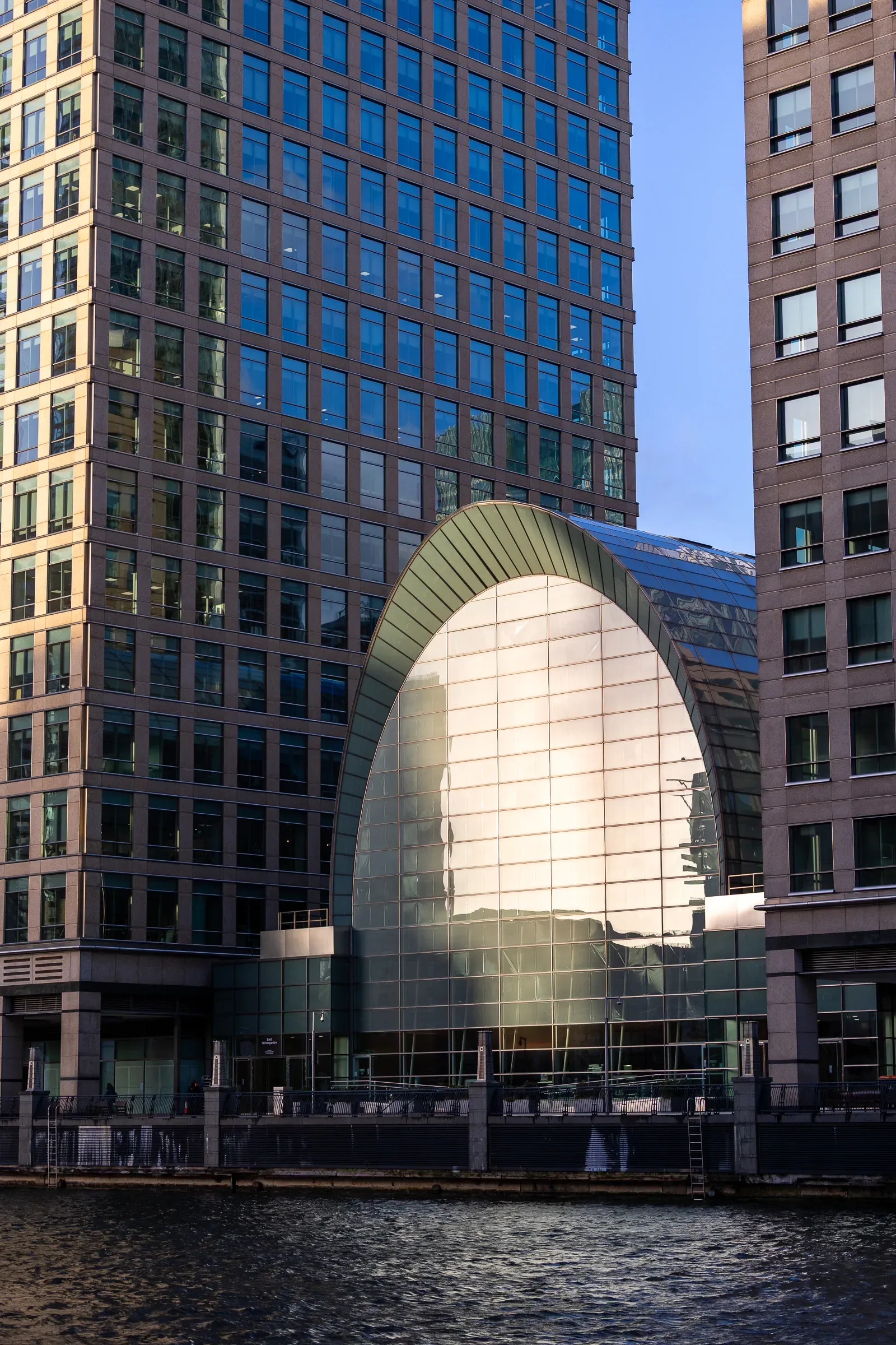 A glass and steel archway between two office towers glowing gold at sunset, reflected in the water below