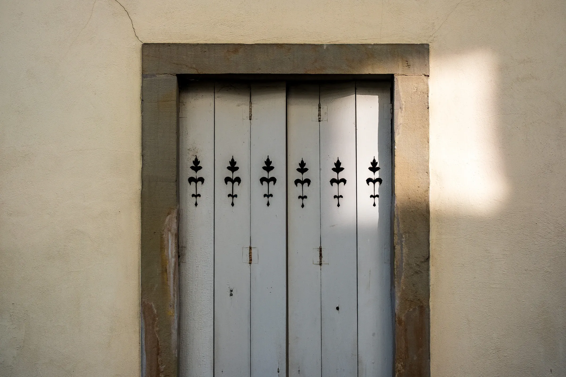A pale wooden door with wrought-iron fleur-de-lis fittings, set into a stone frame.