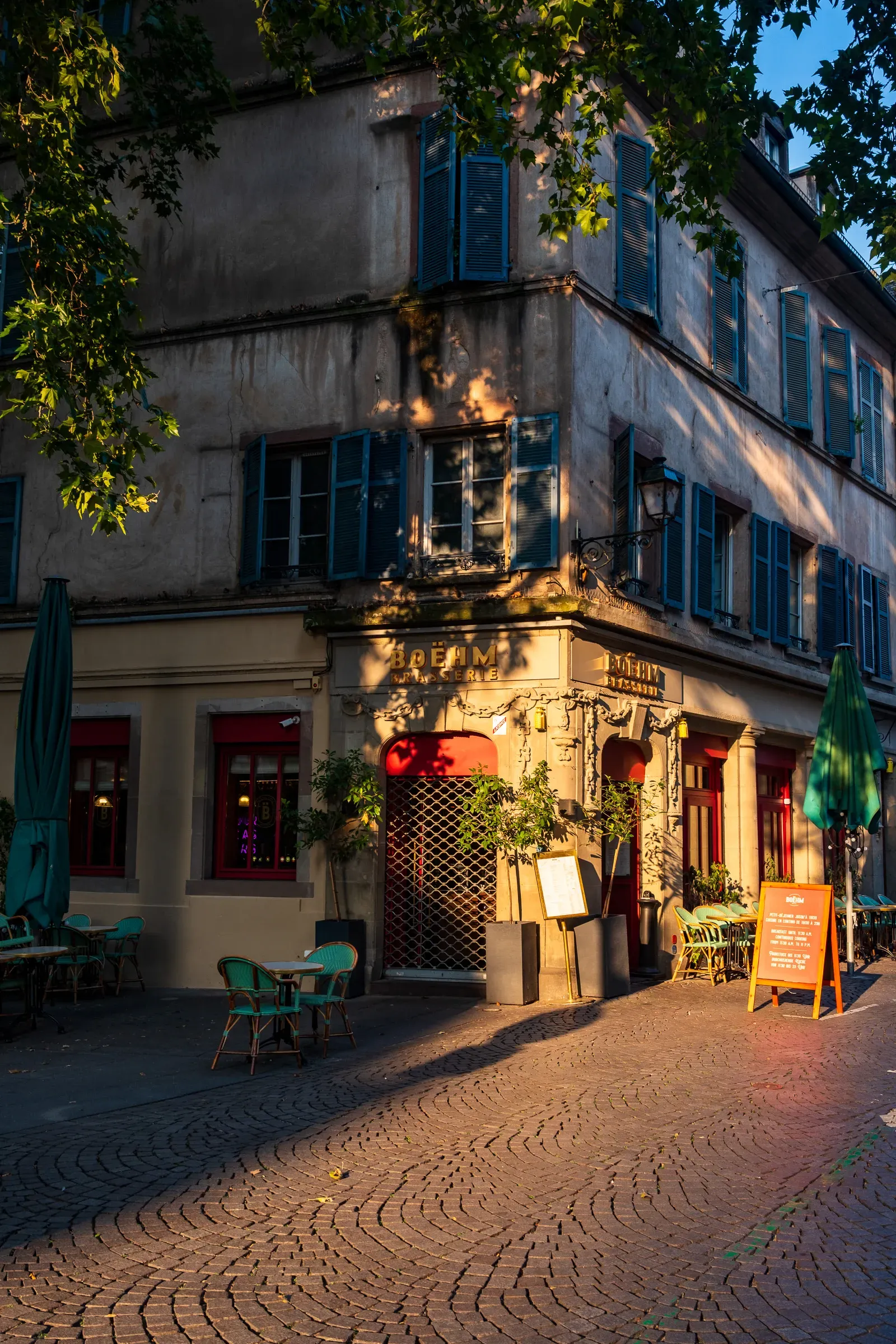 A corner restaurant terrace with green chairs and umbrellas on a cobblestone street, lit by late-afternoon sun.
