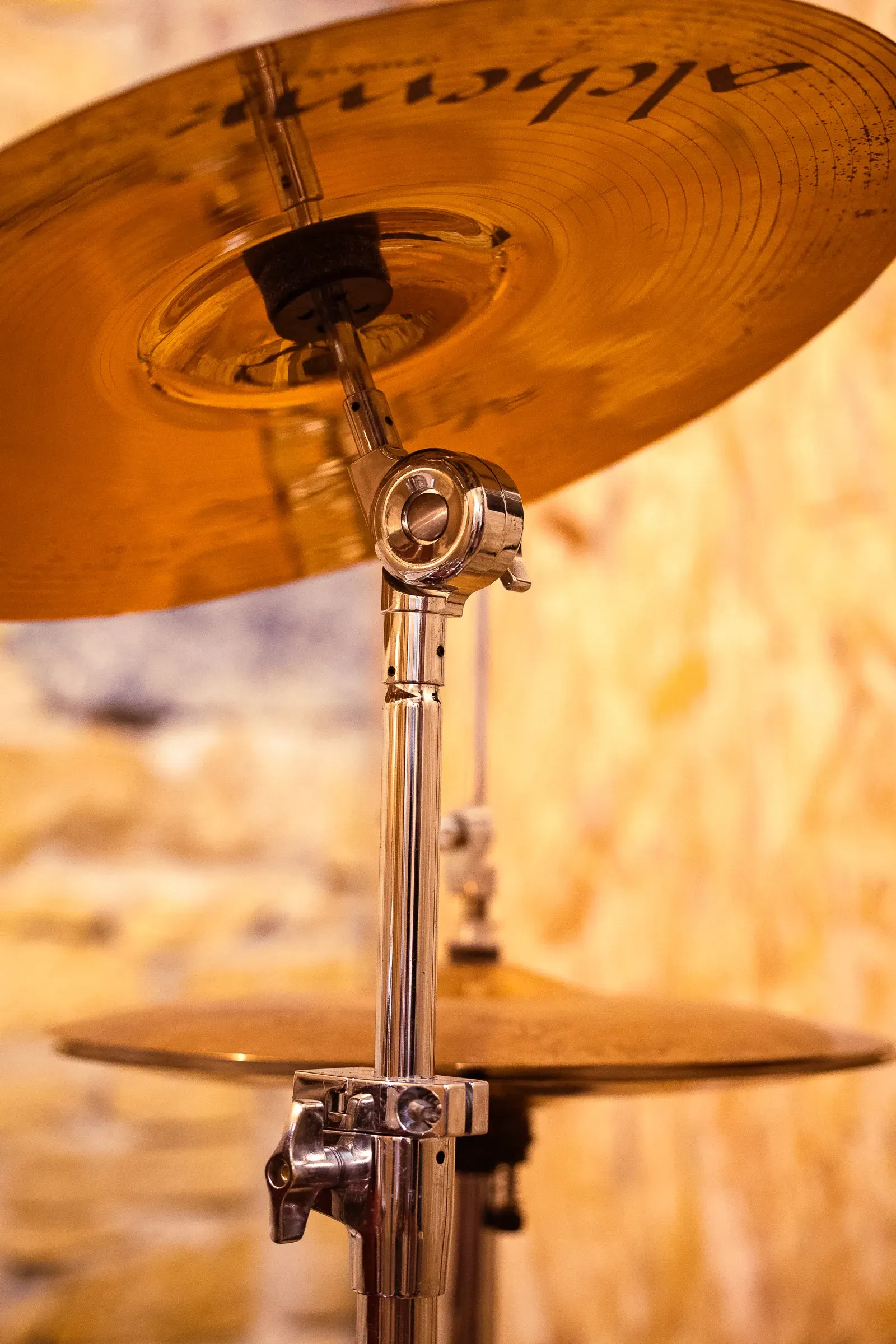 Alchemist hi-hat stand close-up — chrome tilter rod and two cymbals stacked, warm blurred background, Lyon