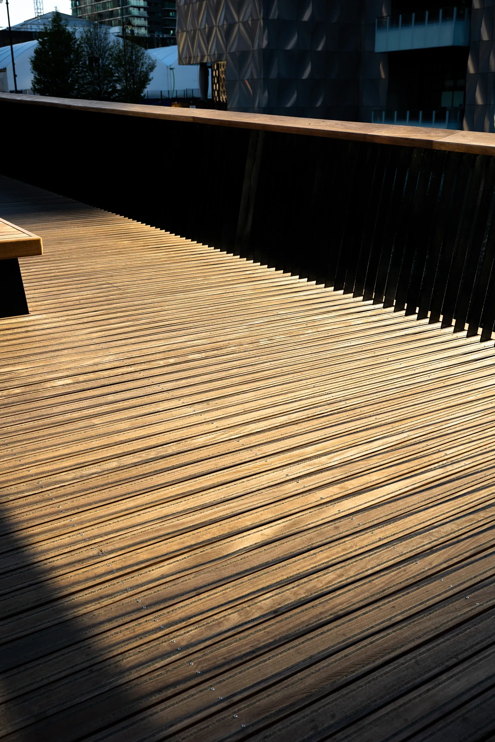A wooden boardwalk in raking evening light, diagonal shadows from a slatted railing, a patterned building behind