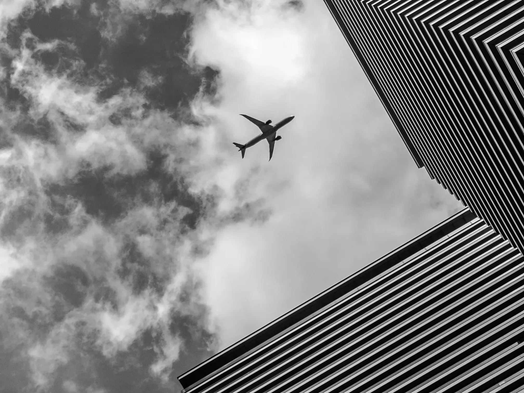 An airplane crossing the sky directly above two glass skyscrapers, seen from street level, black and white