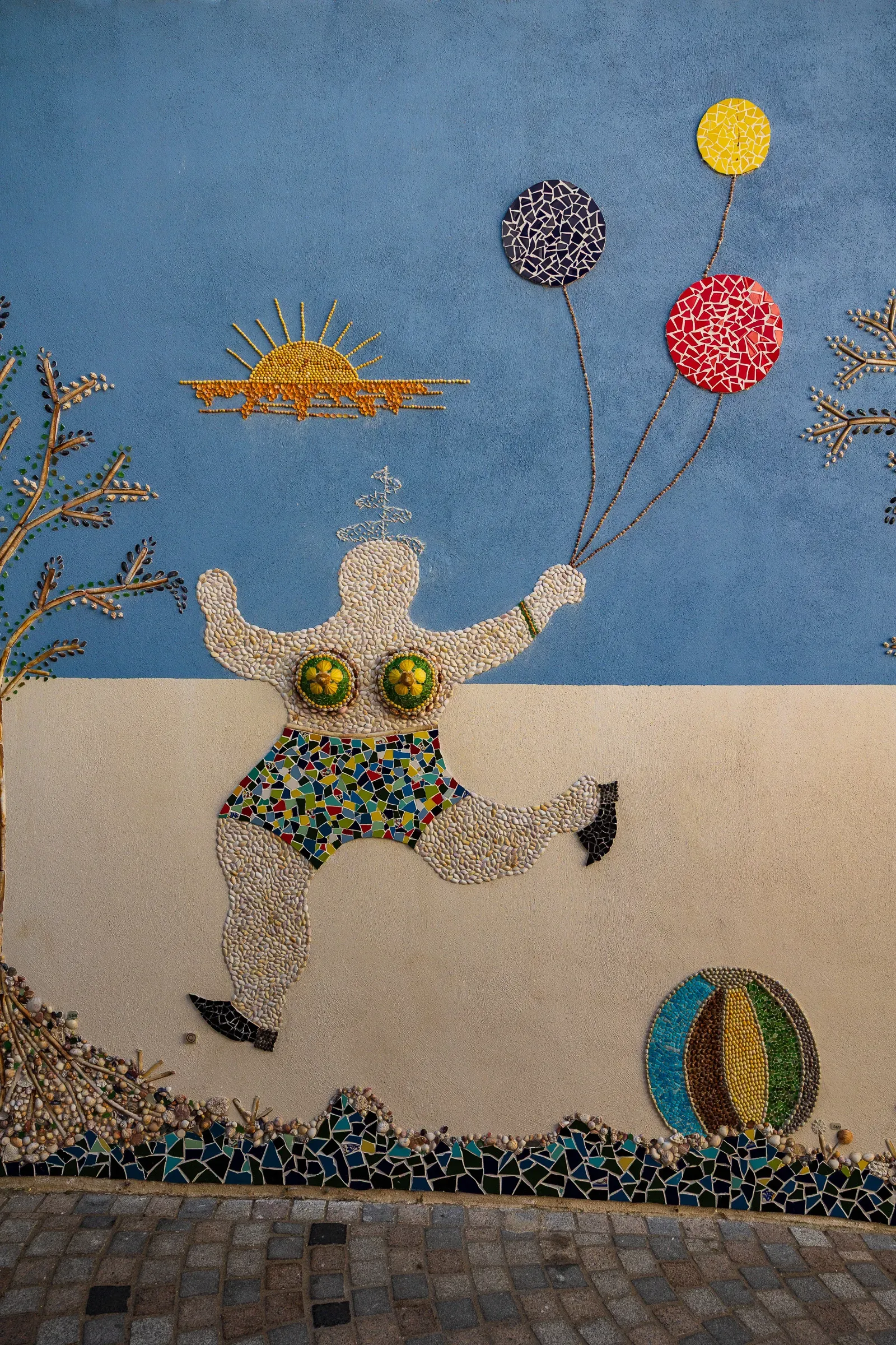 Large mosaic of a woman in a swimsuit holding balloons against a painted blue wall, a beach ball at her feet