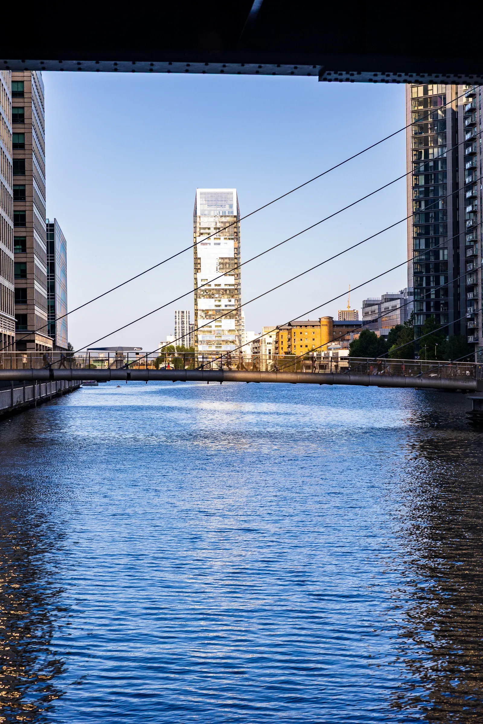 A white skyscraper framed by the suspension cables of a pedestrian bridge over the water, evening light