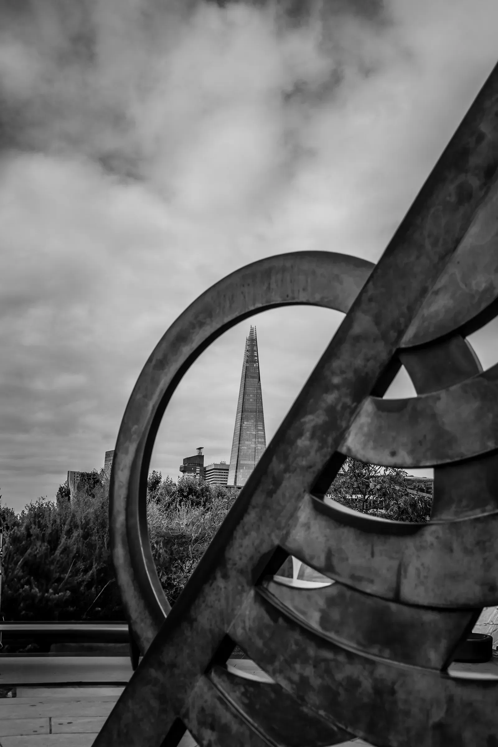 The Shard seen through a circular metal sculpture, London skyline and trees below, black and white