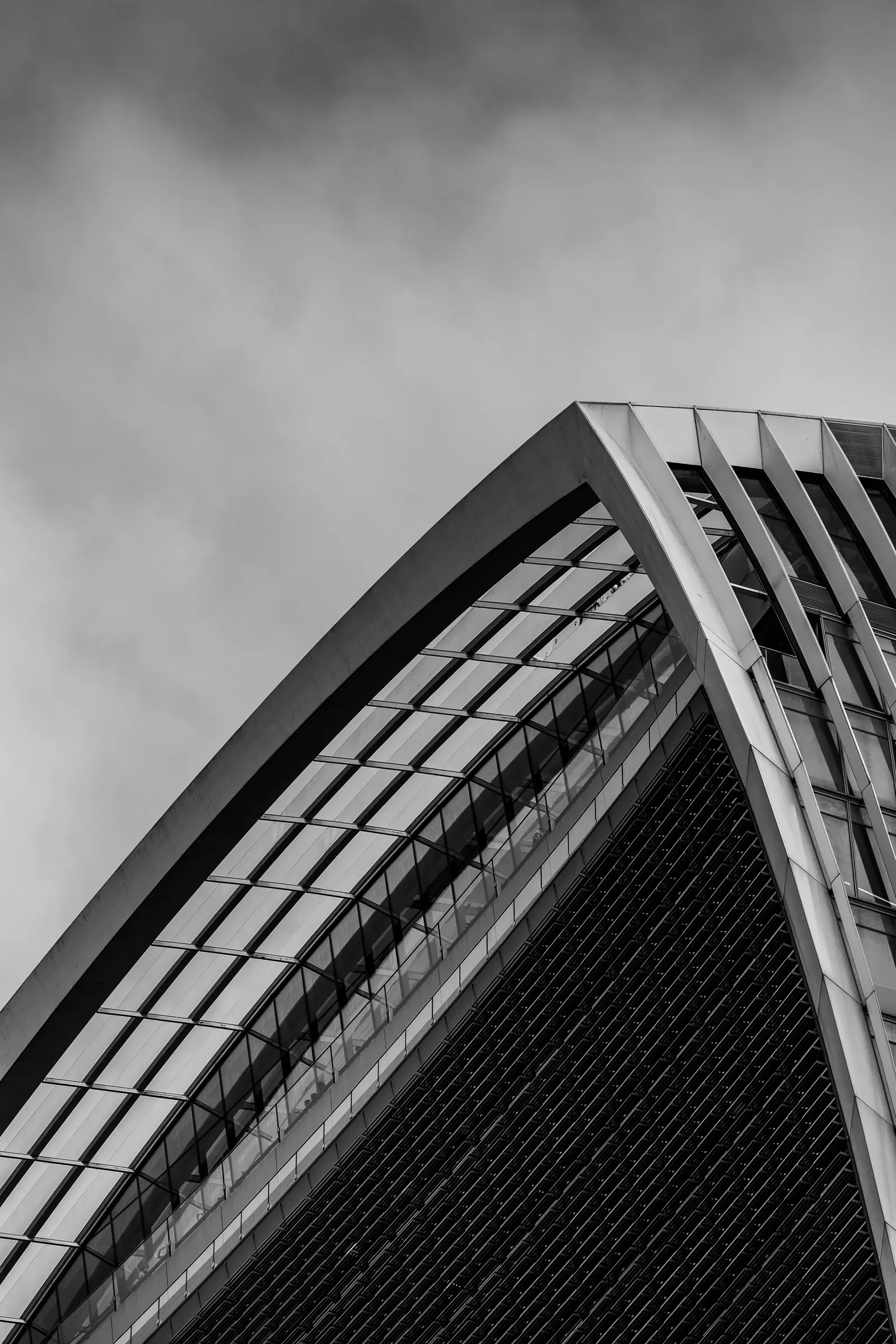 Looking up at the sharp glass and steel edge of a building's canopy against a cloudy sky, black and white