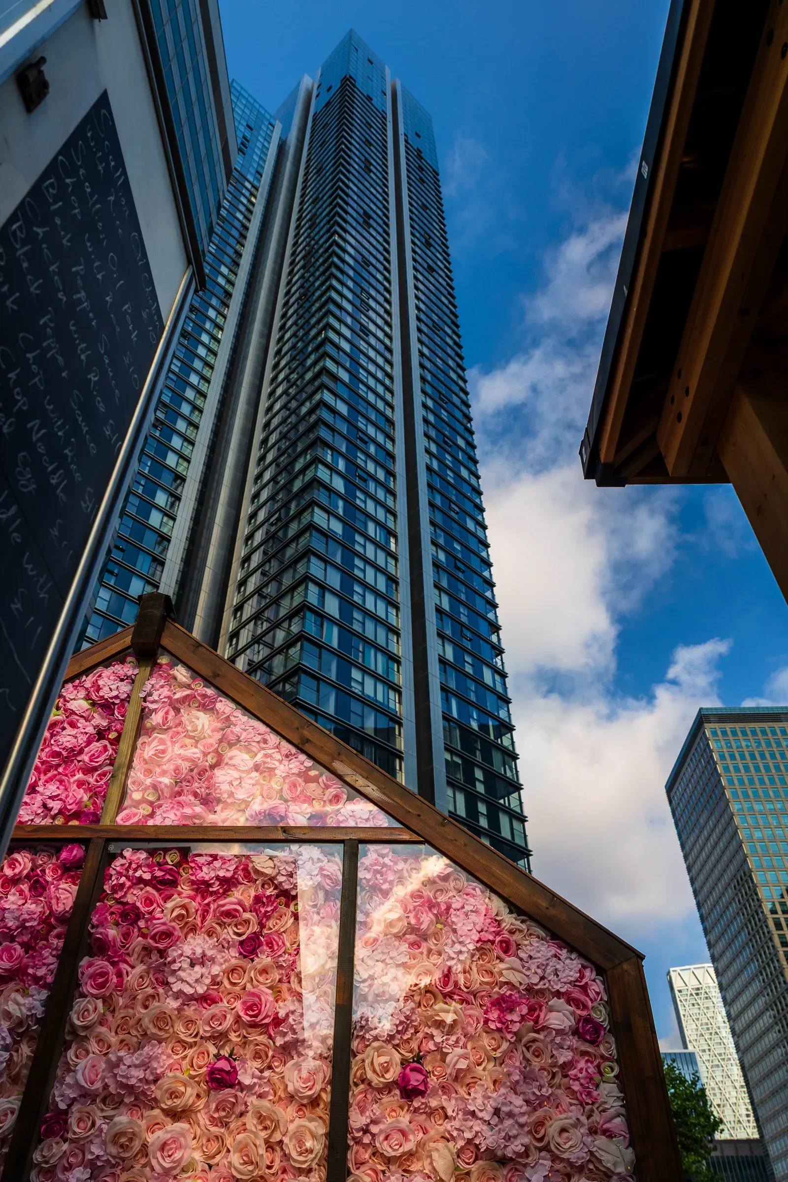 A glass residential tower rising behind a wall of artificial pink roses and a chalkboard menu