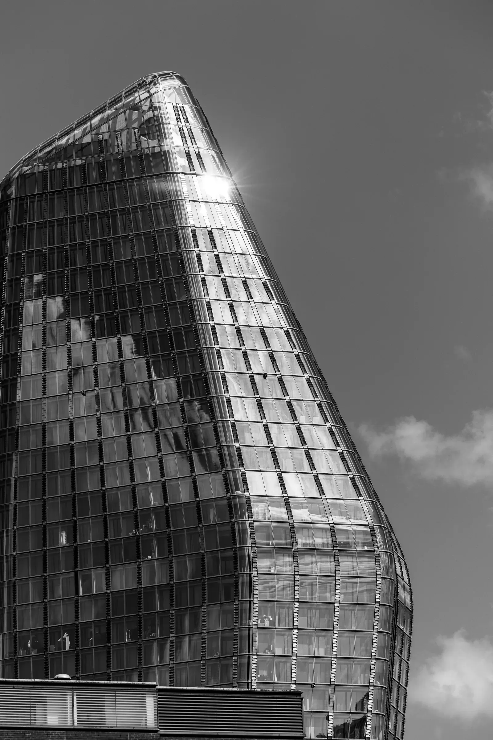 Sun flare on the curved glass facade of a skyscraper, its rounded corner catching the light, black and white
