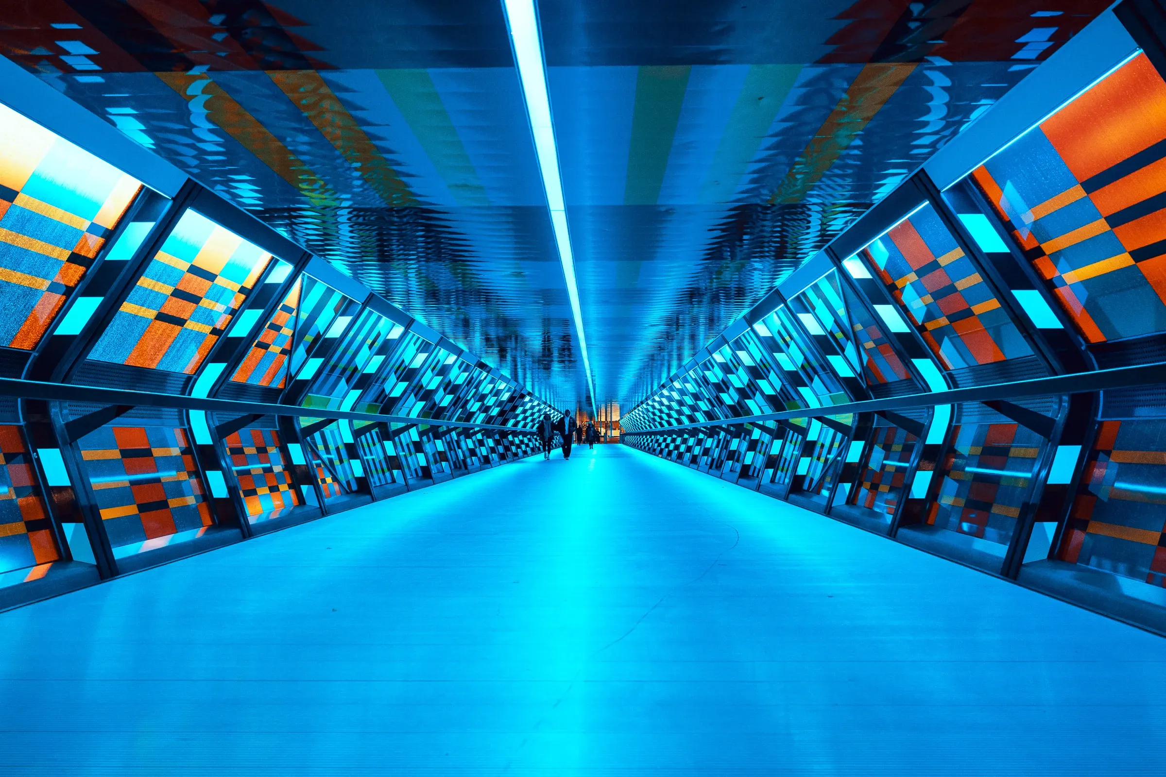 A long pedestrian corridor lined with colorful pixel-patterned LED panels in blue, orange, and green, the perspective vanishing to a distant point — Canary Wharf