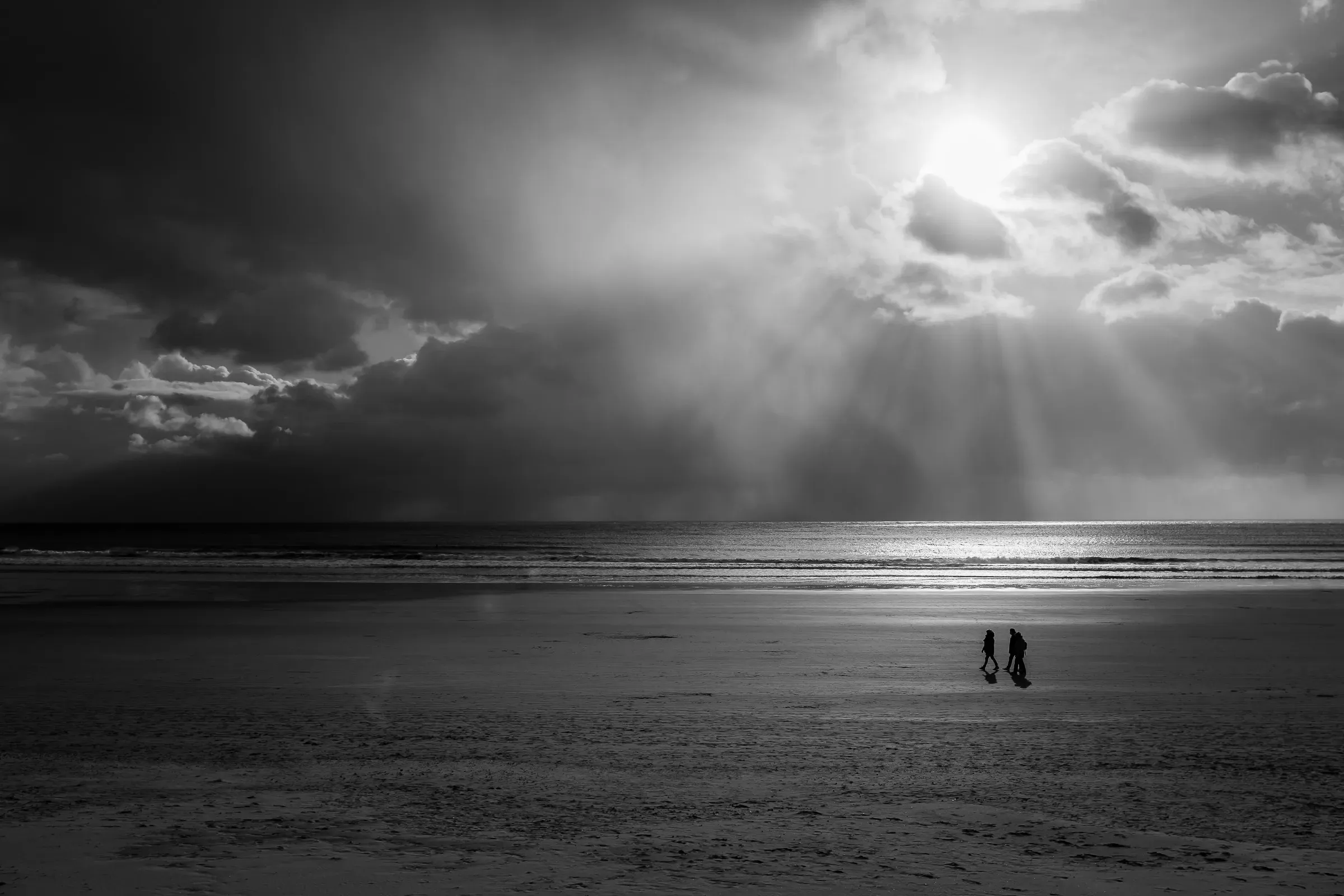 Two silhouetted figures walking a vast empty beach, sunrays breaking through a dramatic storm sky above the sea, light pooling on wet sand