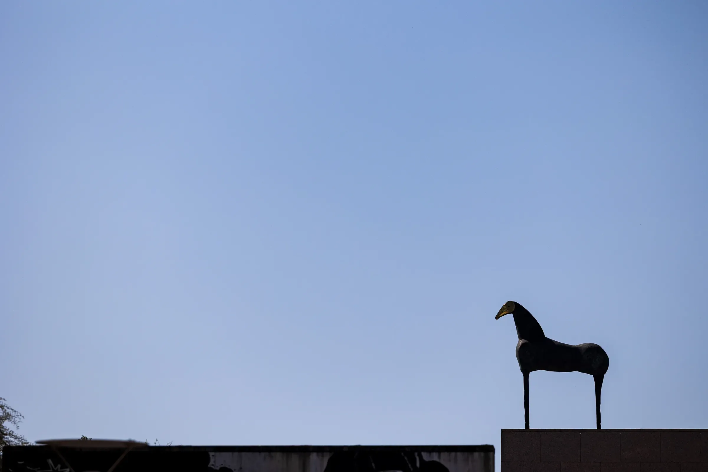 Silhouette of a horse sculpture on a rooftop ledge against a plain blue sky.