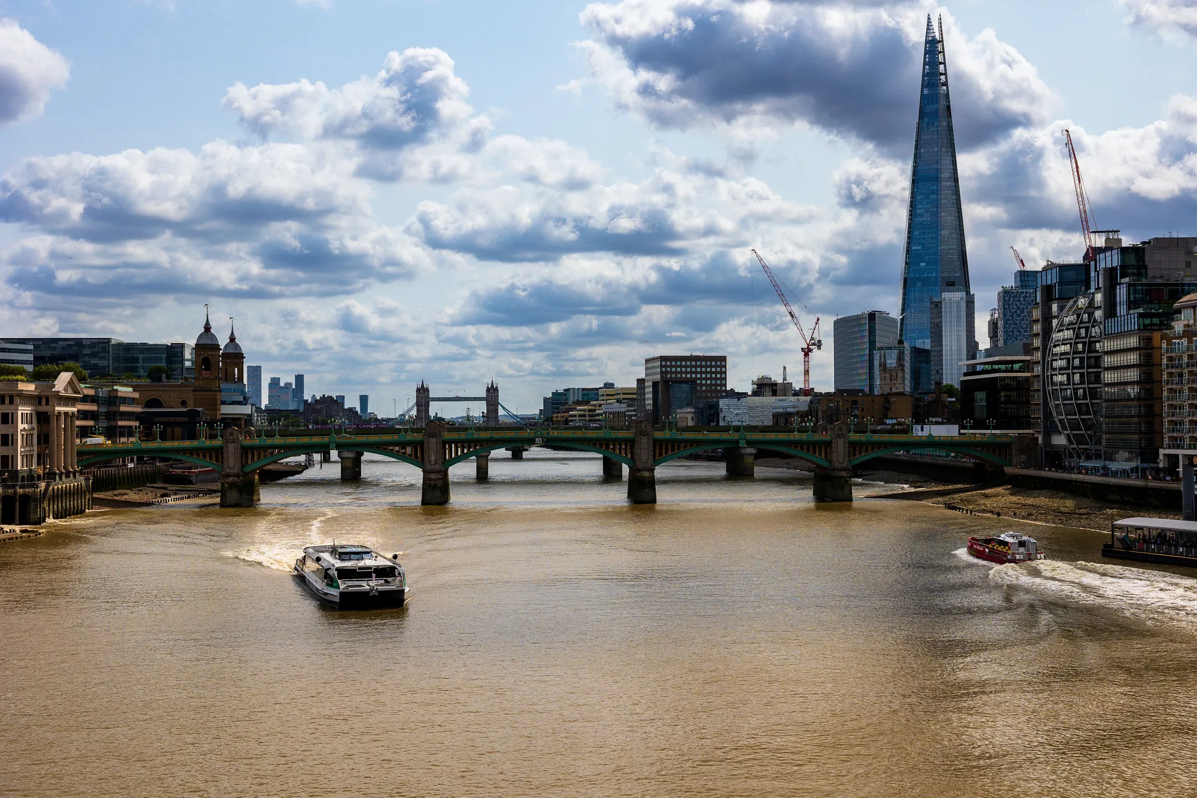 The Shard and Tower Bridge across the Thames, a green railway bridge and passenger boats in the foreground