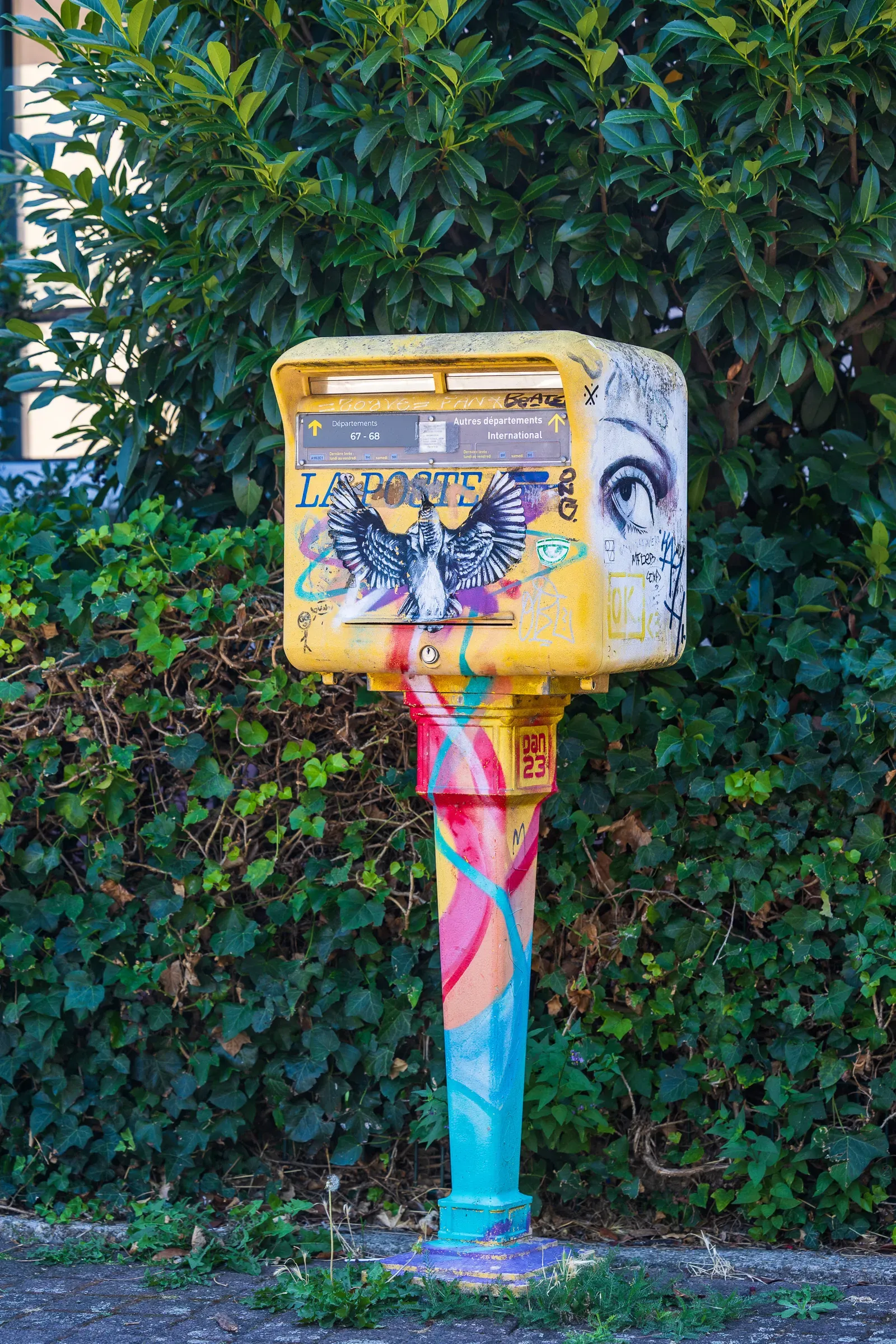 A postbox covered in graffiti and pink-and-blue paint, in front of a hedge.