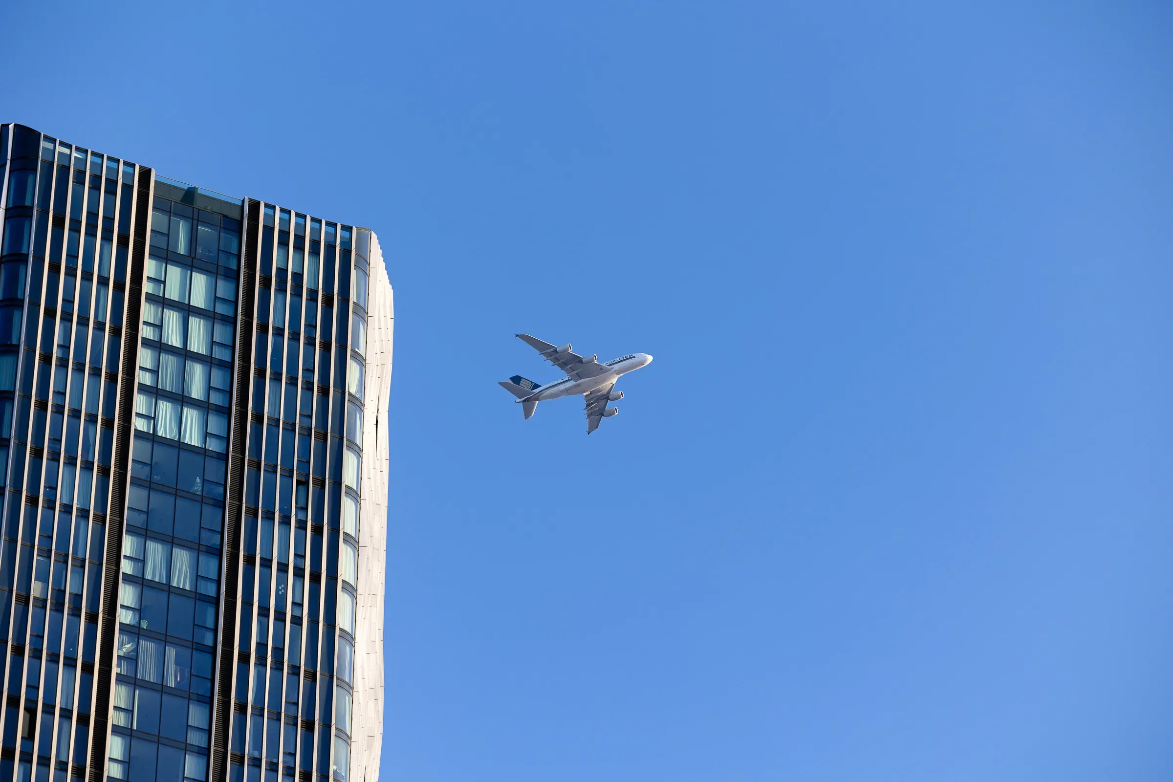 A wide-body aircraft crossing above a glass tower block against deep blue sky, Canary Wharf