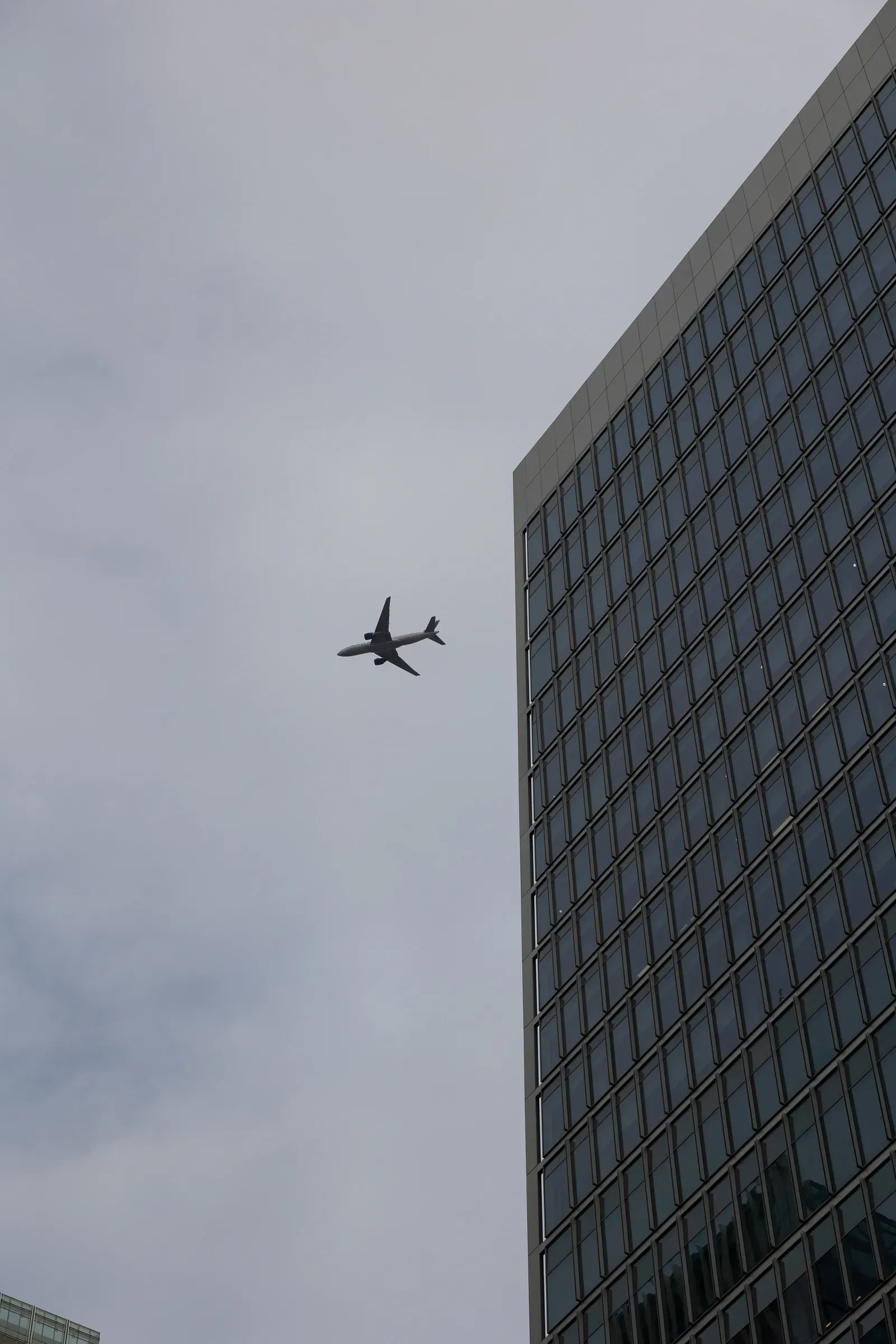 A plane silhouetted against an overcast sky beside the sharp corner of a glass office tower