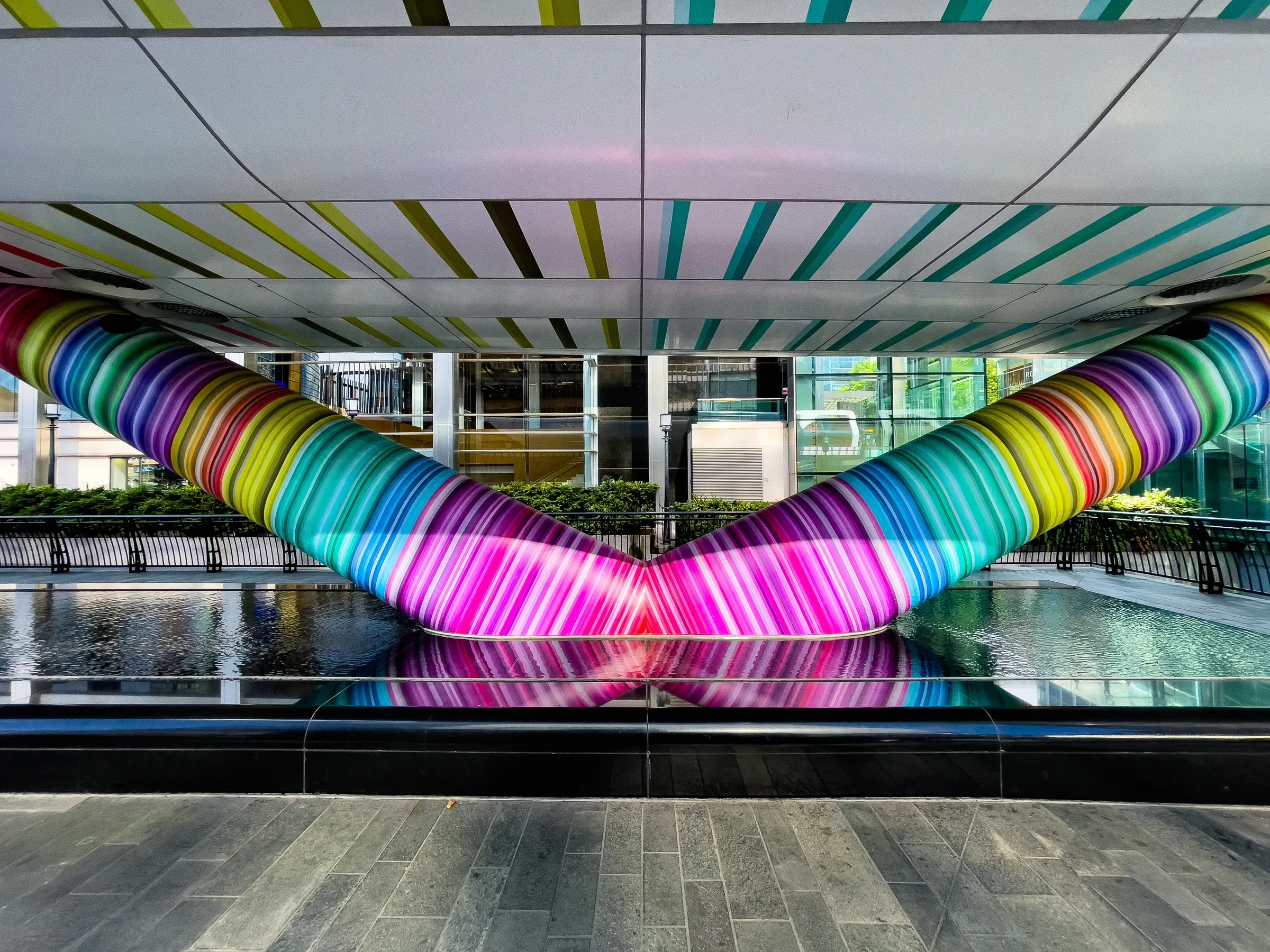 A rainbow-striped tunnel installation reflected symmetrically in a shallow pool beneath a striped ceiling