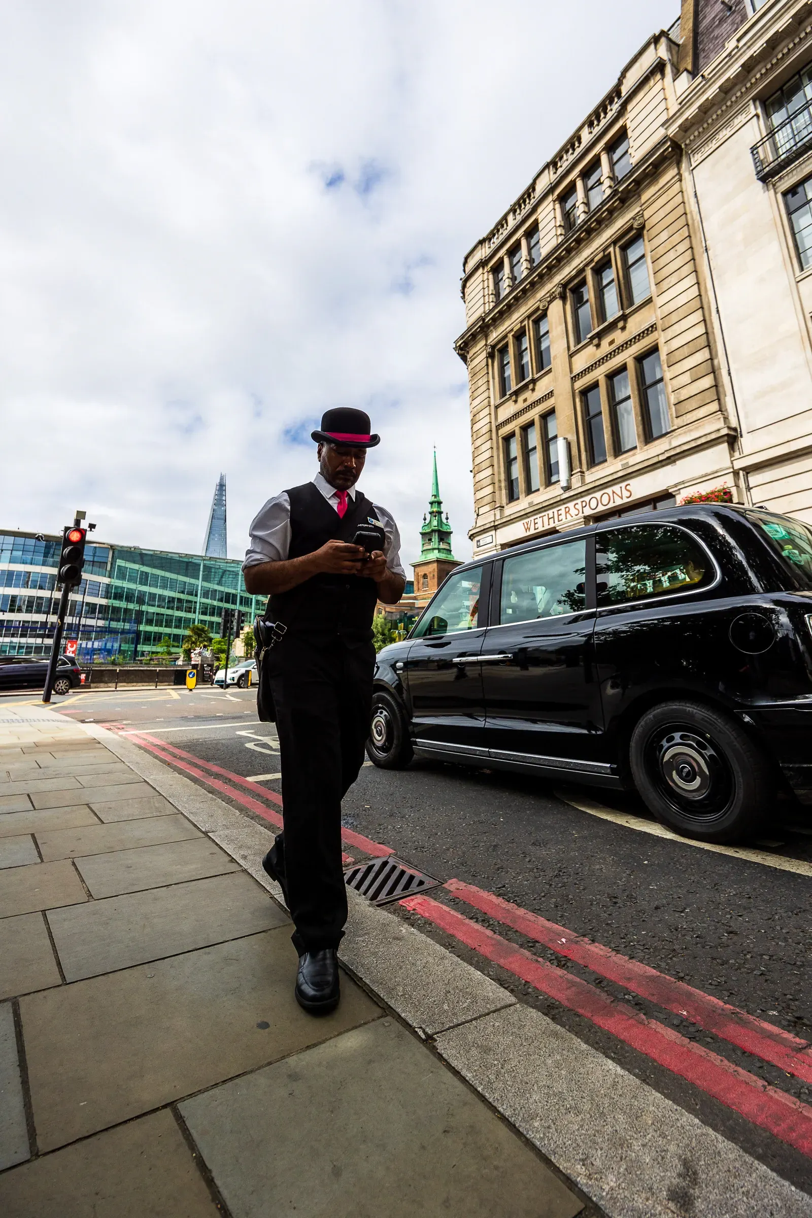 A man in a bowler hat and pink tie checks his phone on a City of London pavement, a black cab and the Shard behind