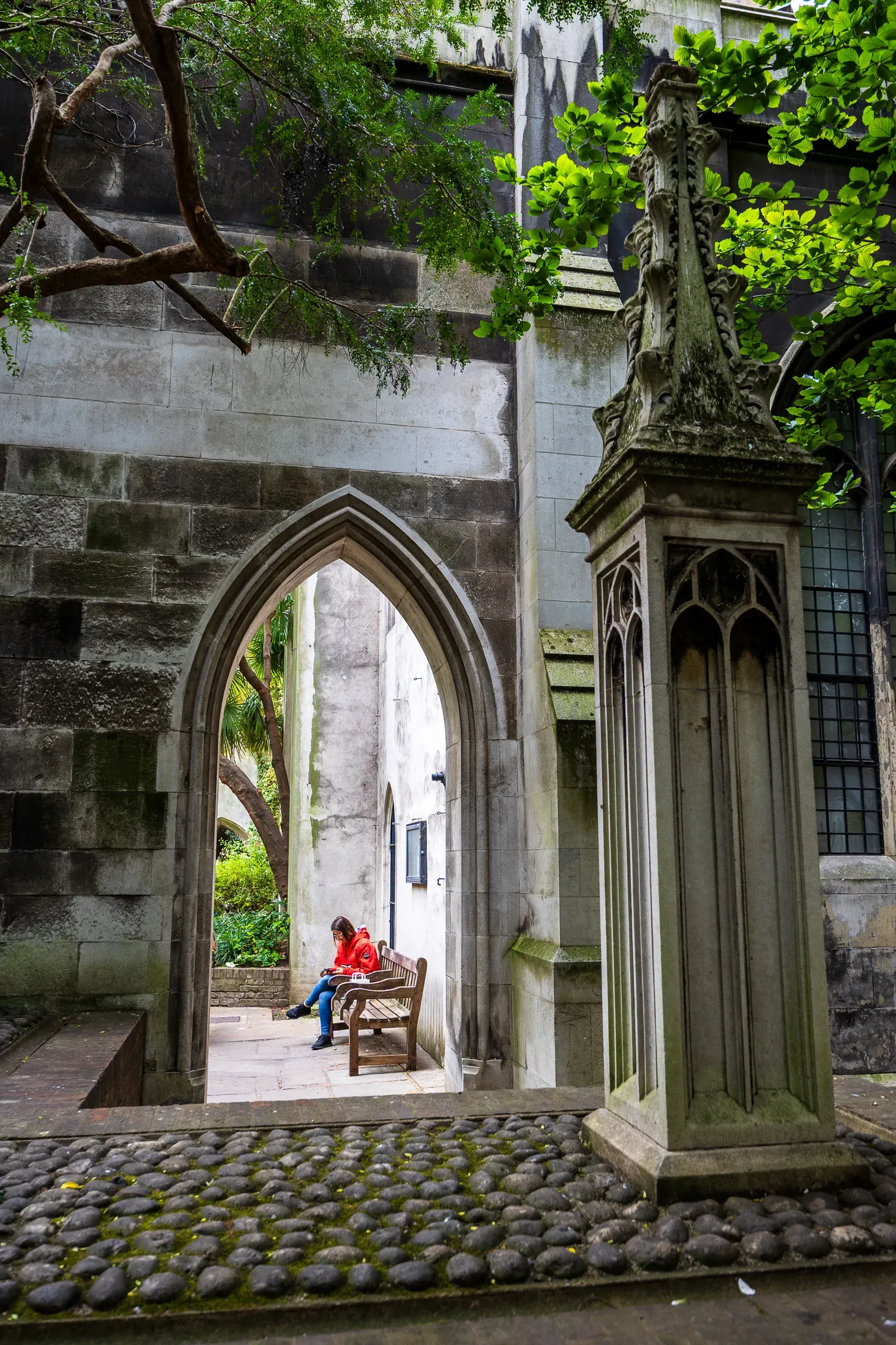 A woman in a red coat reads on a bench, framed through a Gothic stone archway in a ruined churchyard garden