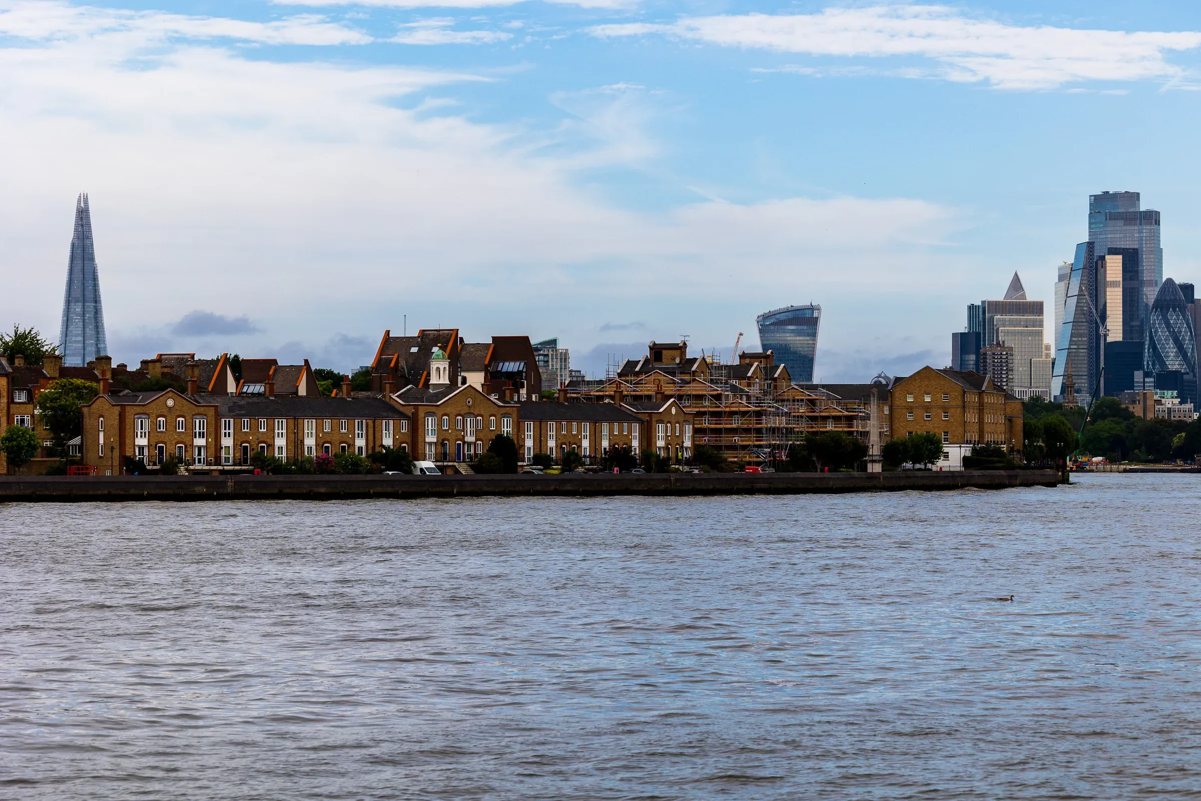 A row of converted wharf houses along the Thames, the City skyline — the Shard, the Walkie-Talkie, the Gherkin — behind