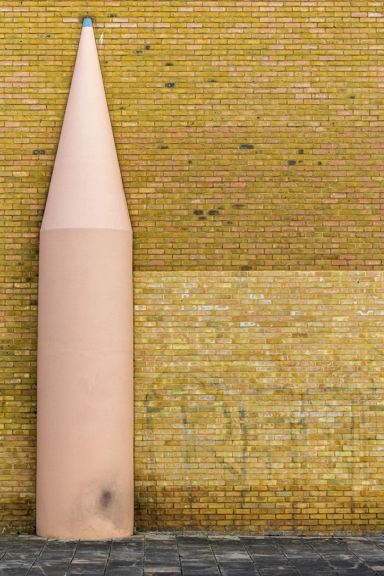 A giant pink pencil sculpture leaning against a yellow brick wall